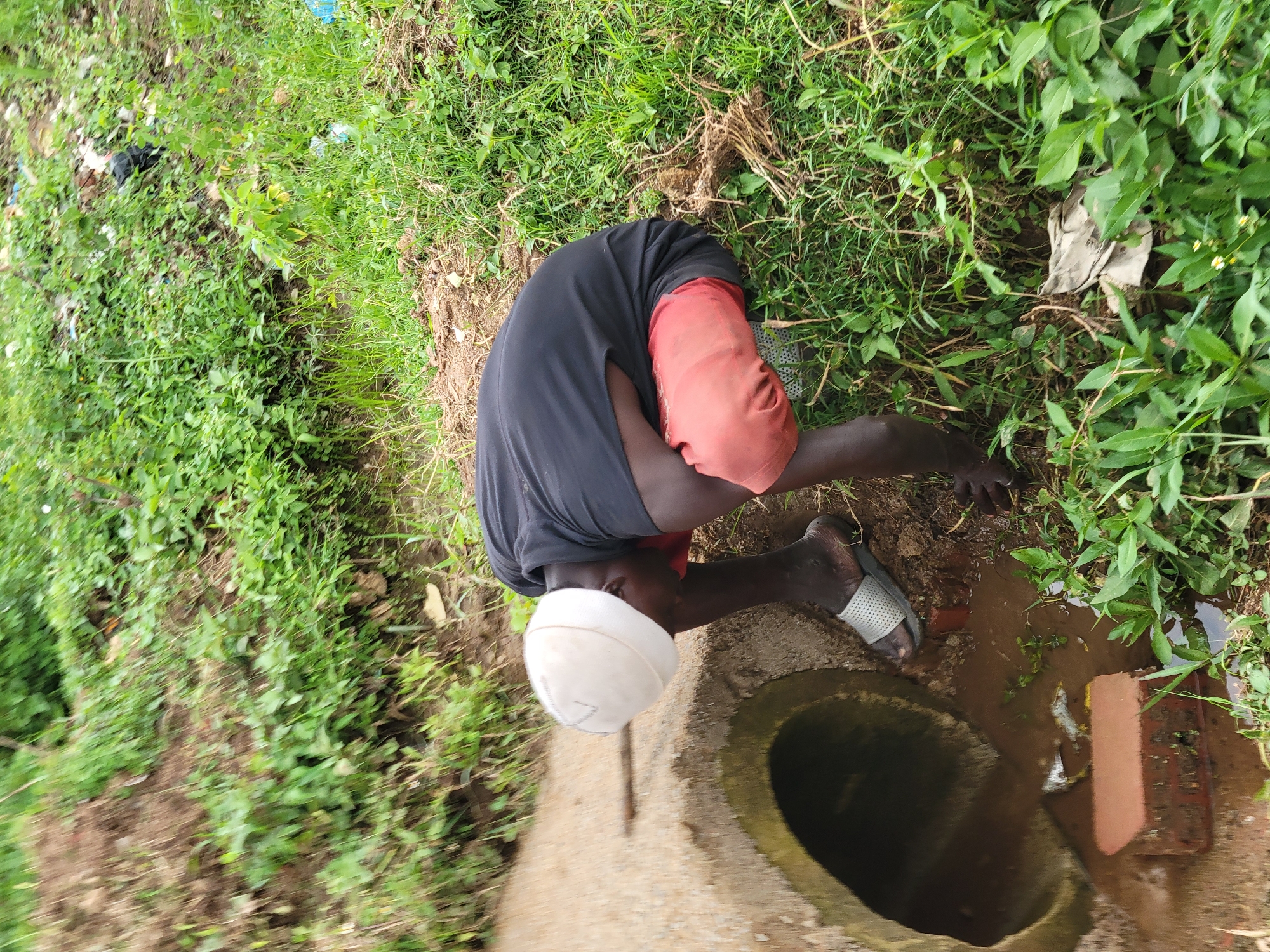 In Barakin Lerawi, Nigeria, a dark-skinned man wearing a black t-shirt, red shorts, a white cap, and grey sandals is crouched by the opening of a concrete culvert. Water flows out of the culvert, and a brick sits in the muddy water. The surrounding area is overgrown with green grass and other vegetation. In the background, there is debris, including plastic bags and what appears to be a black trash bag. The man is reaching towards the culvert opening.