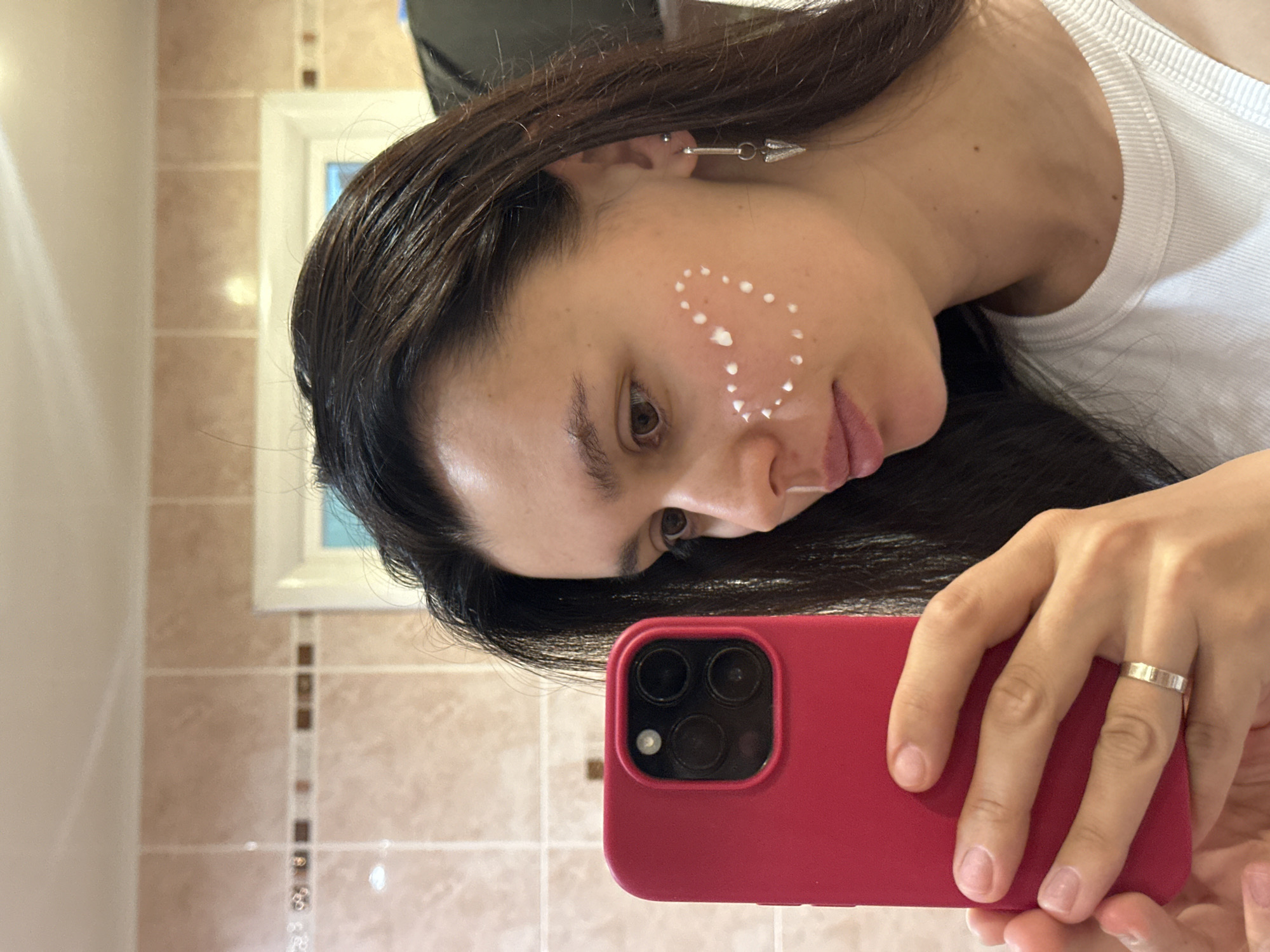 A person with dark hair is taking a selfie in a bathroom. They are holding a red smartphone with three camera lenses. The person has a white cream arranged in a heart shape on their cheek. They are wearing a white ribbed tank top and an earring in the shape of an arrow. The bathroom features beige and brown tiled walls and a window with a white frame. The location is not discernible from the provided image.