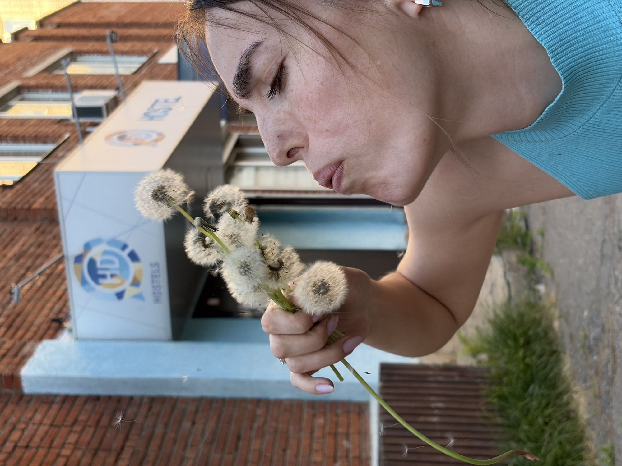 A woman is blowing on a bouquet of dandelions in Izhevsk, Russia. She has fair skin and is wearing a blue top. The background includes a building with "Hostel" displayed and a logo featuring the letters "HD." The building has a red brick facade.