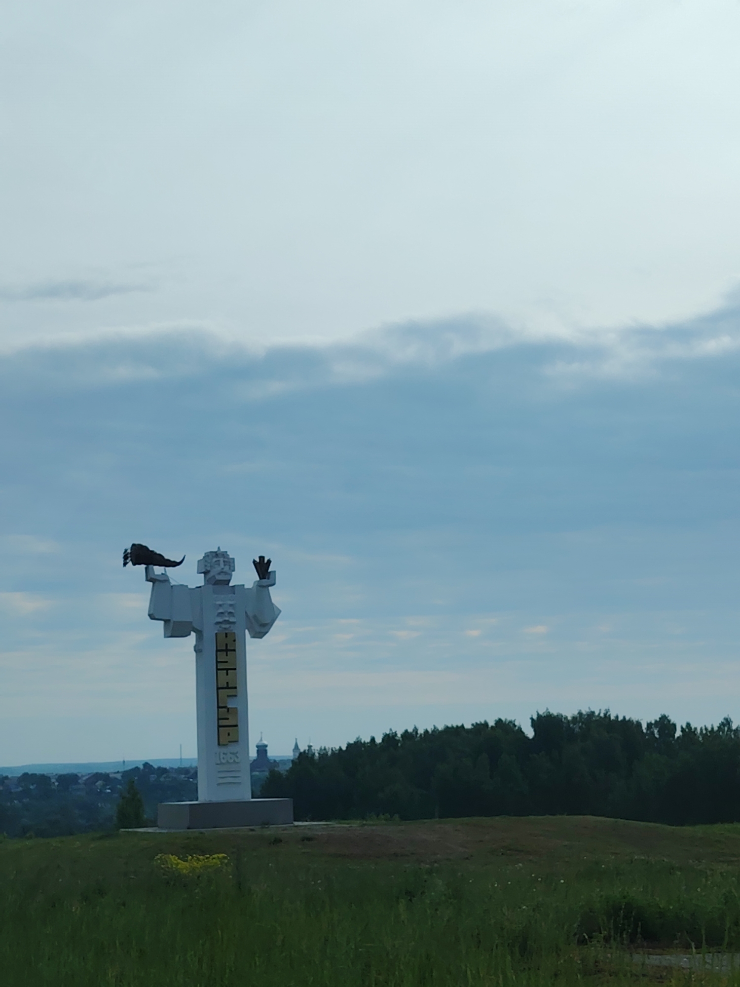 A medium-shot depicts the monument "Пуп Земли" (Navel of the Earth) located in the city of Kungur, Russia. The white monument stands on a grassy hill. The monument is a stylized human figure with its arms raised. The figure is holding a dark object in its left hand. The inscription "1663" can be seen vertically down the front of the monument in yellow letters. In the background, there is a forest line, and beyond it, the city can be seen. The sky is overcast with light blue and grey clouds. The foreground is covered in green grass with some yellow wildflowers.