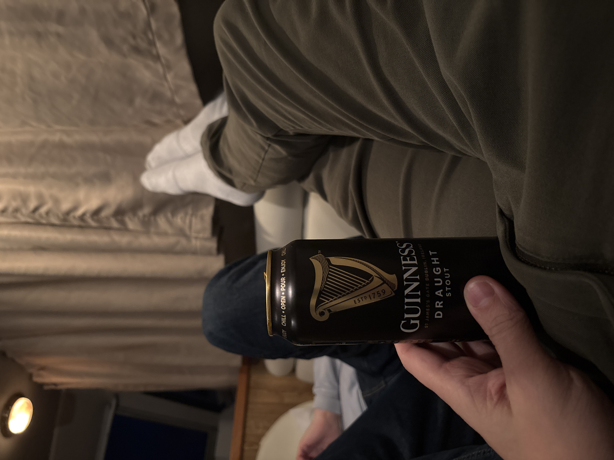 A person is holding a can of Guinness Stout in their right hand while seated. The can is black with gold lettering and the Guinness harp logo. The text on the can reads "GUINNESS", "ST JAMES'S GATE DUBLIN, IRELAND", and "DRAUGHT STOUT". The person is wearing olive green pants on top and blue jeans on the bottom. They also have white socks on. The background includes a curtained window and possibly a bed or seat.
