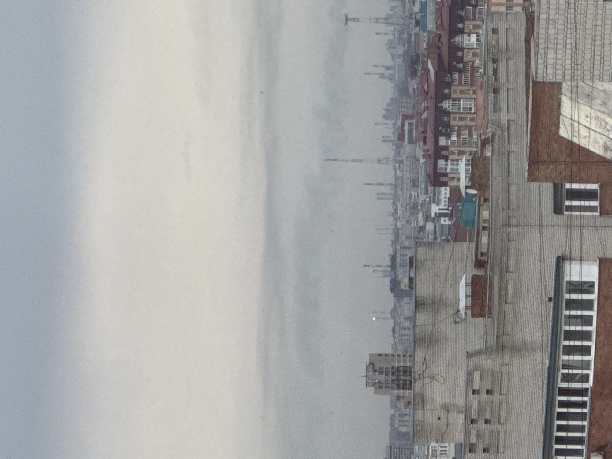An aerial view looking towards the horizon of the city of Unknown, Unknown. On the right side of the image, a row of brick buildings with windows is visible, varying in size. To the left, the buildings fade into the background, obscured by a thick haze that dominates the horizon. Several smokestacks appear in the distance. The sky is overcast and gray, with no visible sun.