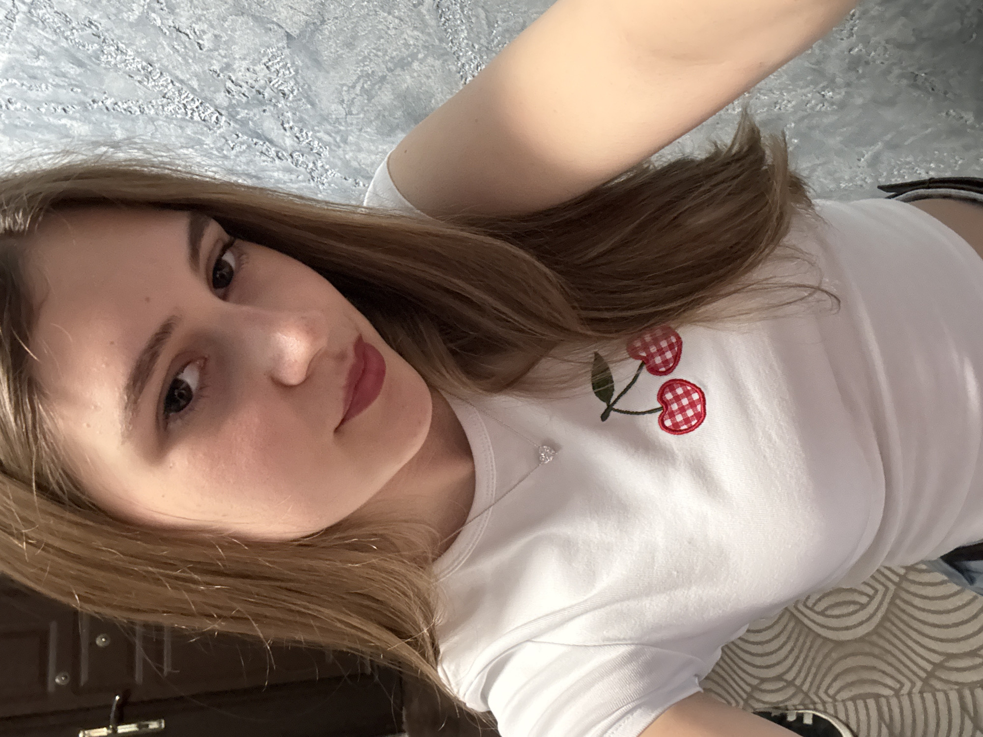 A young woman poses for a selfie. She has fair skin, brown hair, and is wearing a white t-shirt with a red and white cherry design. A silver necklace is visible around her neck. The background shows a textured gray wall and a glimpse of a patterned surface, possibly a bed or sofa. The location in Panagyurishte, Bulgaria is not discernable from the visual information.