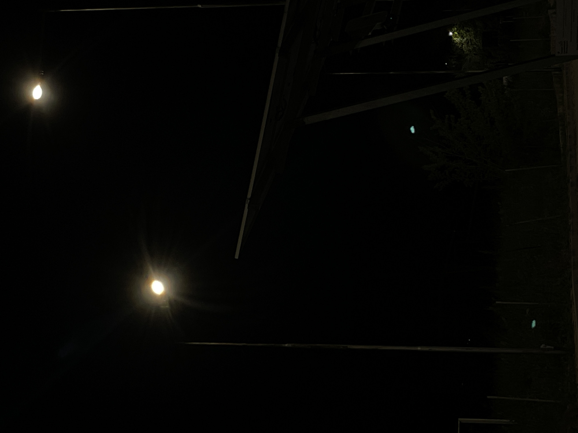 The image depicts a night scene in Byala, Bulgaria. Two illuminated light bulbs are visible in the darkness on the left. Structural elements resembling the framework of a building or awnings are seen in the upper right and middle of the image. Some greenery and additional, fainter lights are visible further to the right. The overall environment is dark with limited lighting.