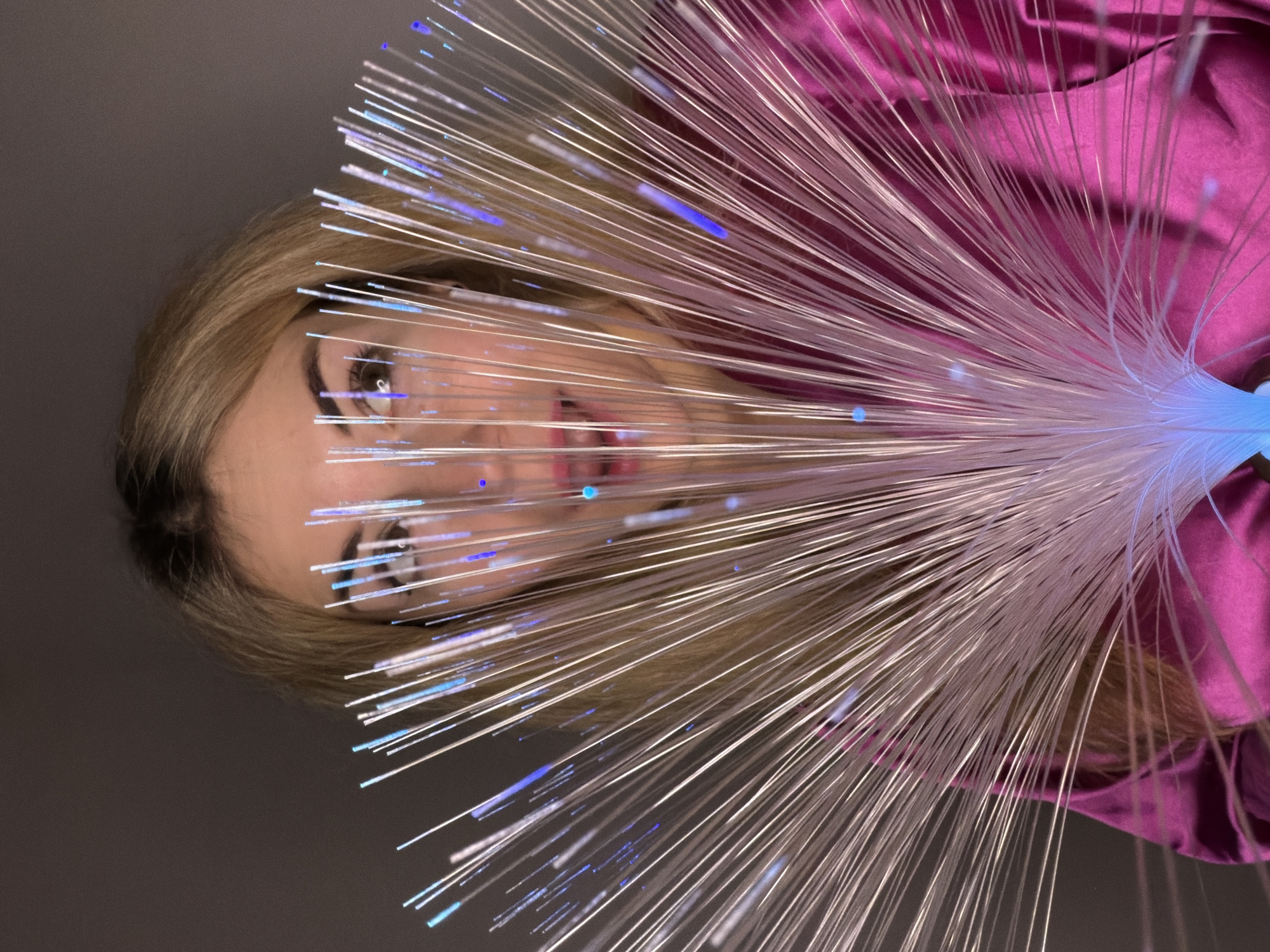 A woman with blonde hair is partially obscured by a fiber optic lamp. The light is emanating from the bottom right corner, obscuring the majority of the woman's face. The fibers spread outwards across the image, some of which are illuminated with blue light. The woman's eyes and mouth are visible through the fibers. She is wearing a magenta-colored top or dress. The background is a plain gray. The image does not provide any indication of it being shot in Budva, Montenegro.