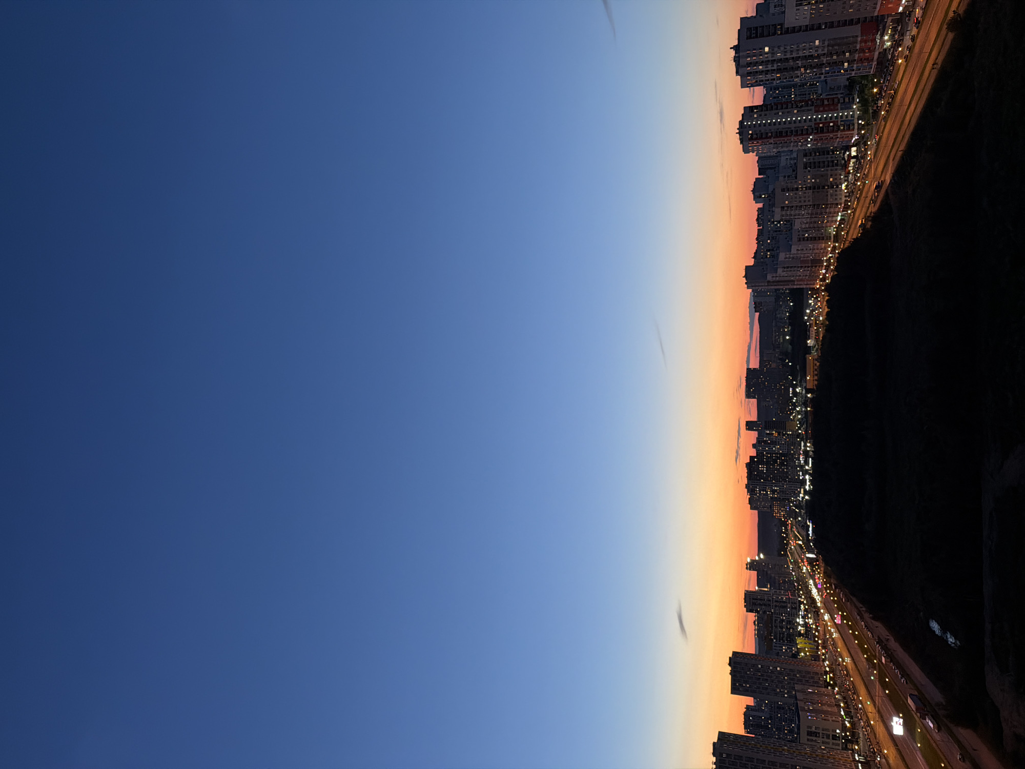 The image captures a cityscape at dusk, likely from a high vantage point. The sky transitions from a deep blue at the top to a warm orange and pink gradient near the horizon, indicating the sun has recently set. A dense cluster of tall, modern apartment buildings dominates the middle ground, their windows illuminated with warm light. A multi-lane highway runs through the city, with streaks of car headlights and taillights suggesting ongoing traffic. The foreground is a dark, silhouetted area of vegetation, possibly a park or undeveloped land. Based on the architectural style and the road infrastructure, this scene is characteristic of the southern residential districts of Yekaterinburg, Russia.