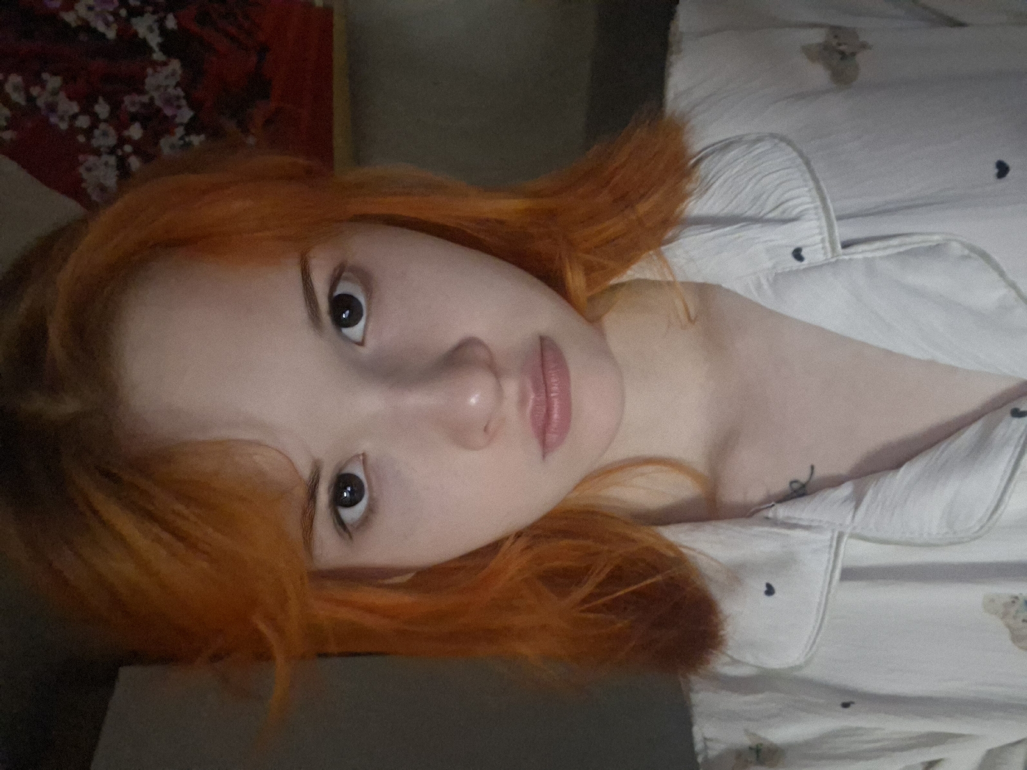 The image displays a close-up, rotated portrait of a young individual with fair skin and vibrant, medium-length orange-red hair. The person is lying down, looking directly at the camera with a neutral expression. One dark eye, the nose, and full, lightly tinted lips are clearly visible, while the other eye is partially obscured by hair.
The individual is wearing a light-colored, possibly white or cream-colored, pajama top. The fabric features small, dark heart-shaped patterns, particularly noticeable on the collar and main body of the shirt. A faint, light brown teddy bear design is also discernible on the fabric in the upper right portion of the garment. A bare shoulder and upper chest are visible, indicating the top has an open neckline or is a button-up shirt worn loosely.
In the background, behind the person's head, there is a dark red surface adorned with a white or light-colored floral or abstract pattern, suggesting a tapestry or artwork. The immediate foreground below the person's face consists of a dark, indistinct surface. The lighting is soft and even, typical of an indoor environment, possibly a bedroom given the casual attire and pose. There is no visible text in the image. The setting for this scene is Chaikovsky, Russia.