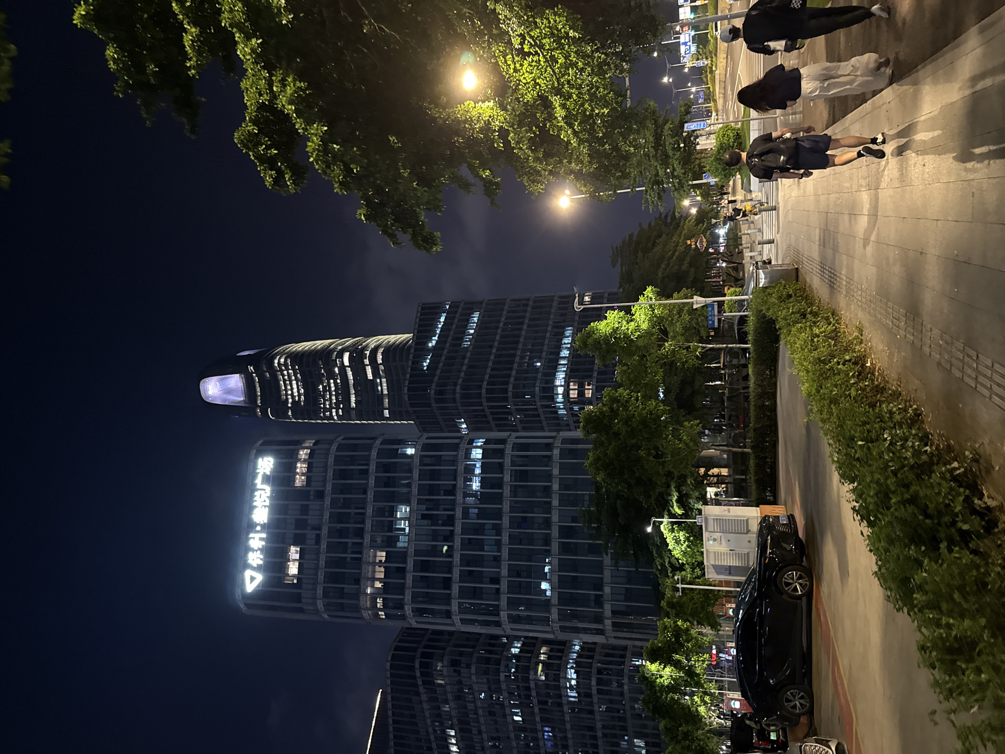 This is a nighttime, low-angle, wide shot of a street scene in Haizhu District, China, featuring modern architecture and pedestrian activity. Dominating the left side of the frame are several glass-facade skyscrapers, illuminated from within and with prominent neon signage on one building displaying "科•盈翠广场" (Ke Ying Cui Plaza). The tallest building on the right has a distinctive illuminated spire with a purple hue. A black sedan is parked on the right side of the street, partially obscured by a white utility box. In the foreground, a paved sidewalk is lined with dense green hedges and trees. Three people are walking away from the camera on the sidewalk: a man in athletic shorts and a dark t-shirt with a backpack, followed by a woman in light-colored pants and a dark t-shirt, and another person in dark clothing. Streetlights cast long shadows across the sidewalk. The road is visible in the background, with streetlights and the glowing windows of buildings stretching into the distance. The sky is dark, indicating it is nighttime, with some faint clouds visible.