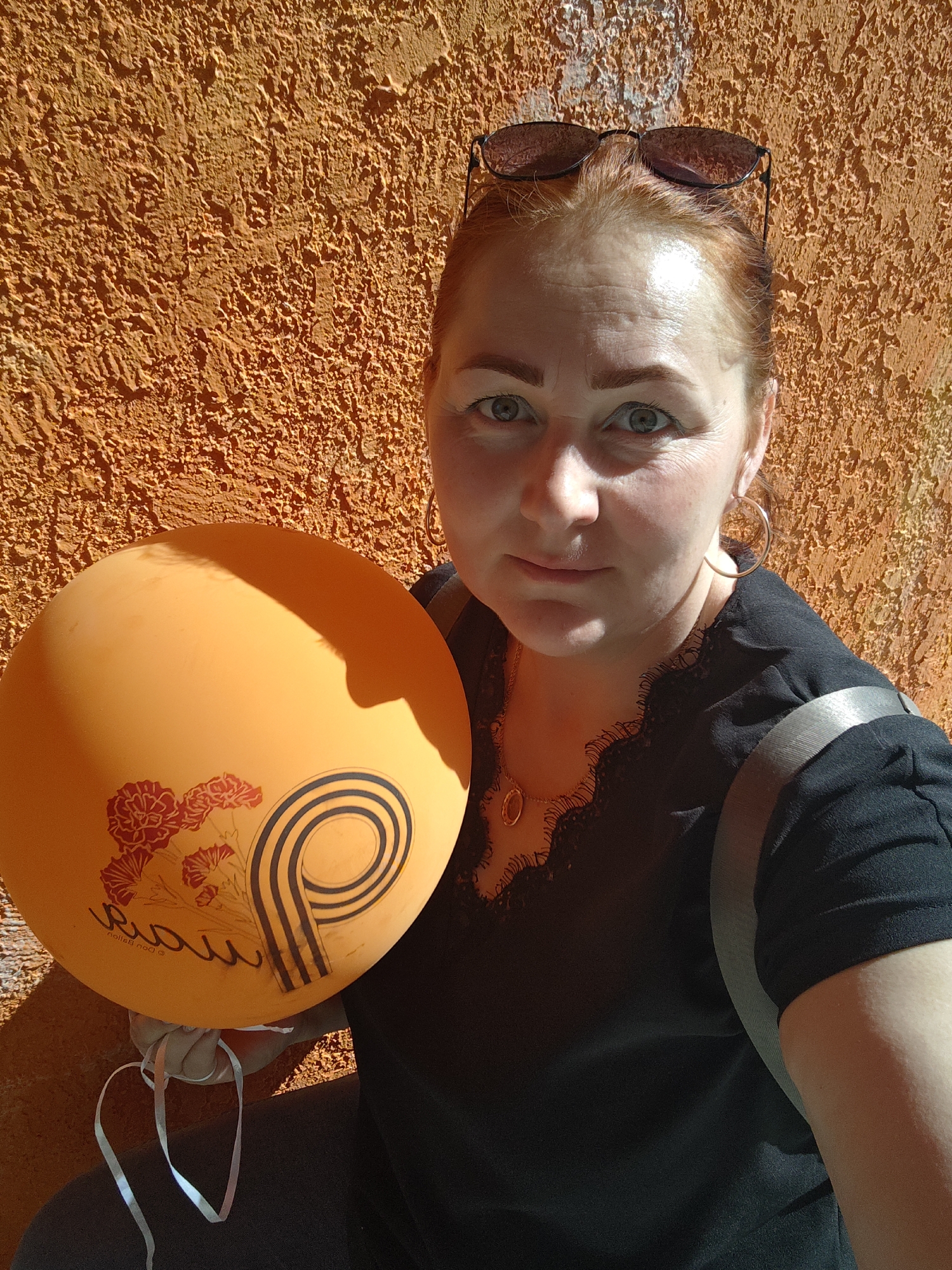 A woman with red hair, fair skin, and sunglasses perched atop her head poses for a selfie. She wears a black shirt with lace detailing around the neckline, gold hoop earrings, and a gold necklace. A silver strap crosses her body. She holds an orange balloon adorned with red flowers and black and white stripes. The background features a textured orange wall. The location is Марковский, Russia, suggested by the writing on the balloon.