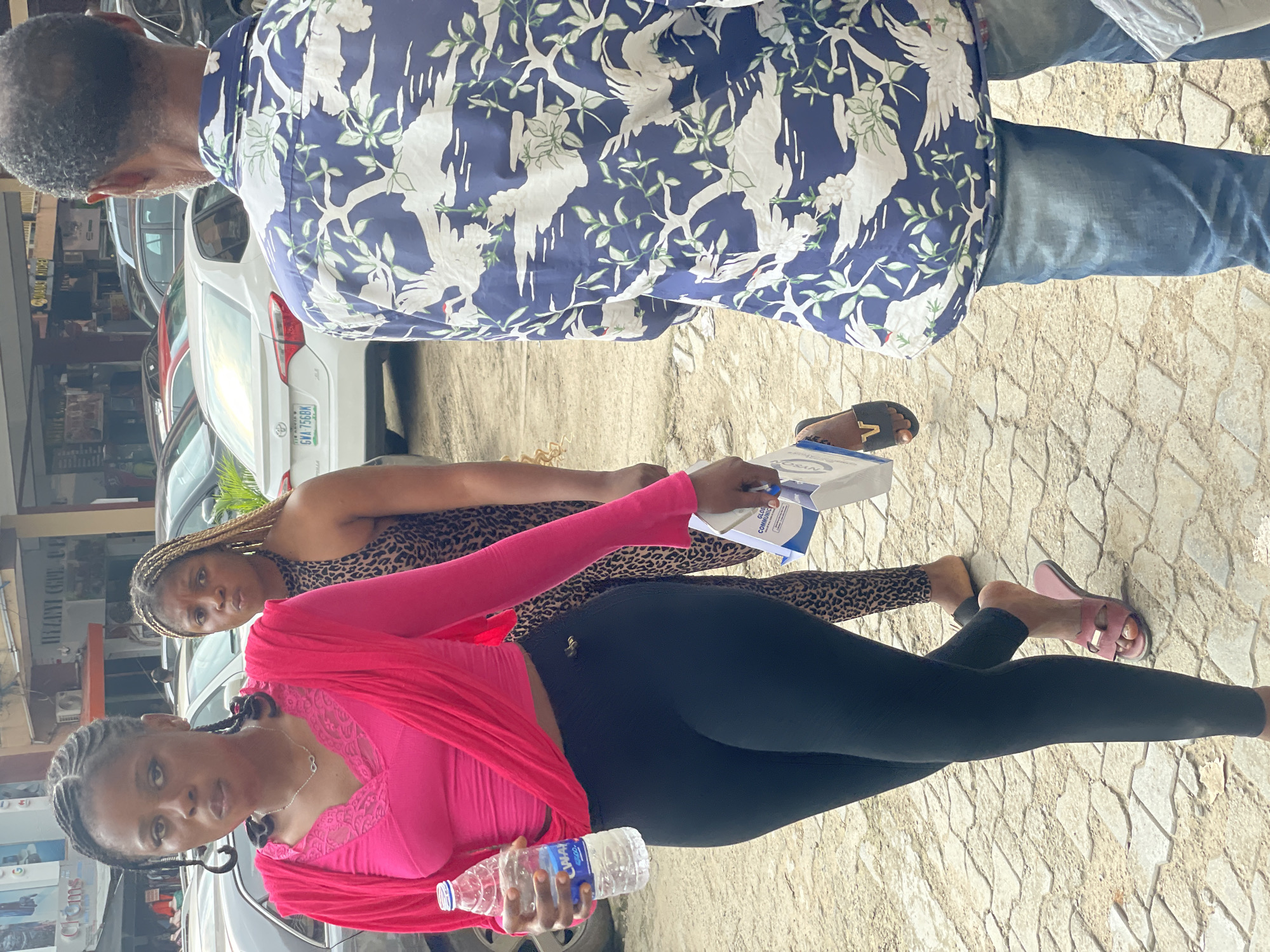 In Abuja, Nigeria, on a paved city street, a young woman in a bright pink long-sleeved top and black leggings is holding a bottle of water. She is positioned in the foreground and appears to be looking towards the camera. Behind her, another woman in a leopard-print top and dark leggings walks away from the camera, carrying a white paper bag. To the right of the frame, a man wearing a blue shirt with a white bird and leaf pattern and blue jeans walks away from the camera, his back is to the viewer. Several cars are parked on the street in the background, including a white Toyota Corolla. The environment suggests a busy urban setting with commercial buildings visible behind the individuals.