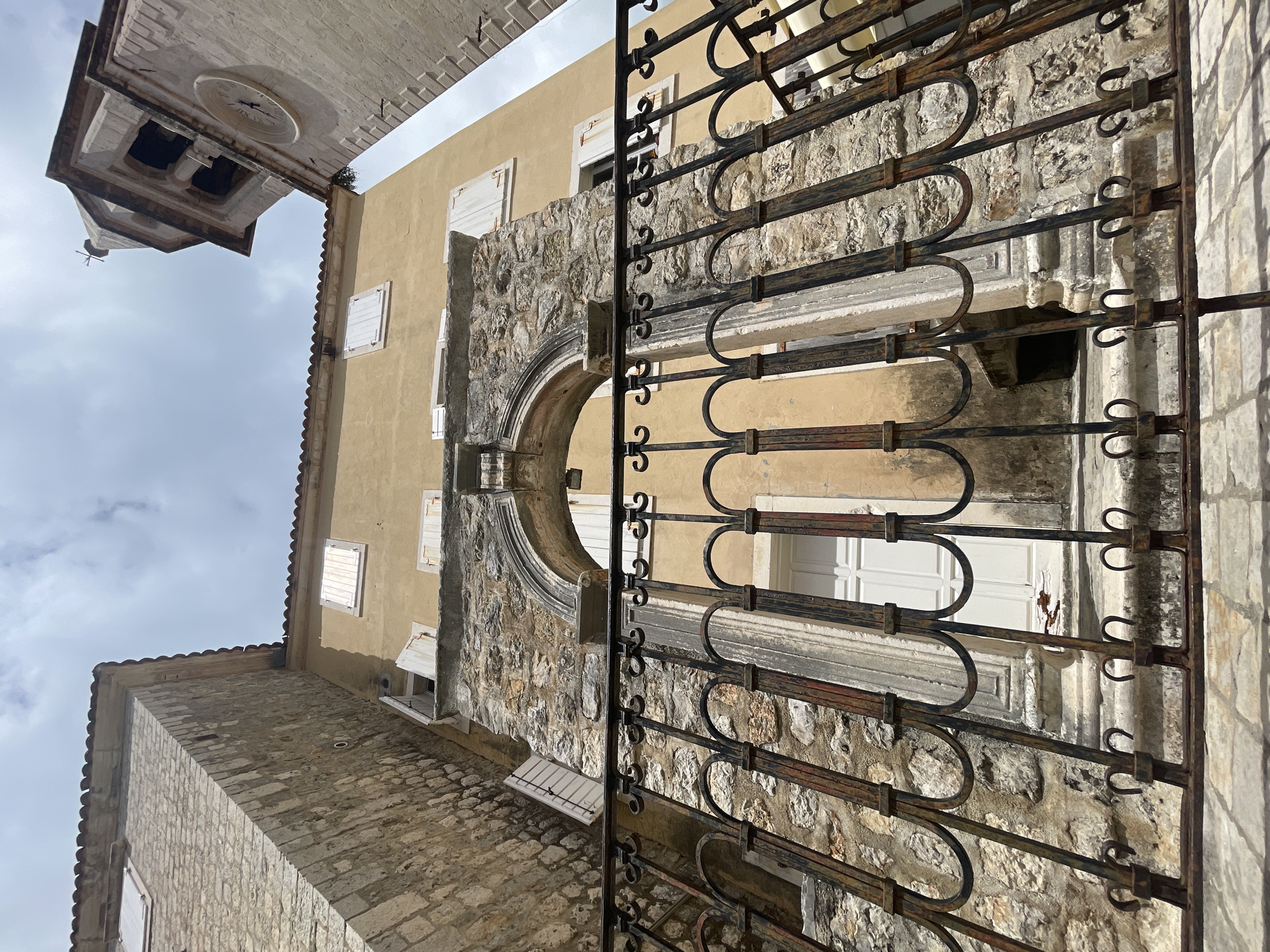 The image depicts a vertical street-level view looking upwards into what appears to be an old European town or city. In the immediate foreground, a weathered and ornate wrought-iron gate or fence runs vertically, partially obscuring the scene beyond. The gate is dark brown and black, with decorative scrollwork that shows signs of rust and age.
Beyond the gate, the scene features several historical buildings constructed from stone and stucco. A light yellow/ochre colored building stands prominently, featuring multiple windows with closed white wooden shutters. Below one of these windows, a small, open, white shuttered box structure is visible. Adjacent to this building, a robust stone wall with an arched opening and a decorative stone balustrade or archway adds architectural interest. The ground level below is paved with rough, uneven cobblestones.
In the upper left portion of the image (when oriented correctly), a stone bell tower with a clock face rises against an overcast sky. The clock, which is light-colored, shows the time to be approximately 10:45 or 11:45. The bell tower has distinct architectural details, including what appears to be a cross on its pinnacle, though it is small and partially obscured by the sky. The sky is largely covered with light gray clouds, suggesting an overcast day. No people or specific activities are visible in the static architectural scene.