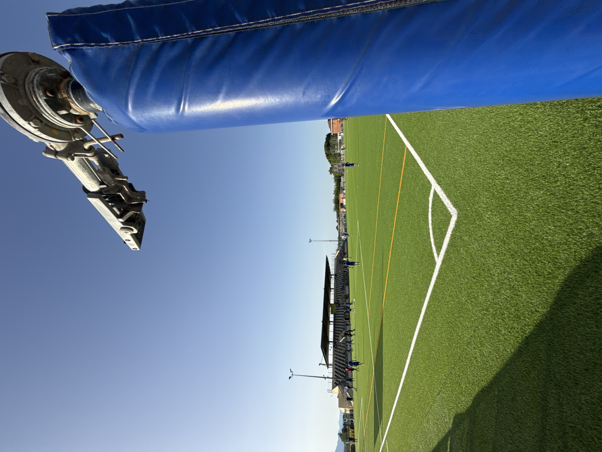 The image depicts an artificial turf soccer field in Vidreres, Spain. A blue padded post is in the foreground, partially obscuring the view of the field and a grandstand in the background. Several individuals, appearing to be soccer players, are scattered across the field, some engaged in activity. The sky is clear and blue, suggesting a sunny day. The scene is set in a sports facility with lighting poles and surrounding buildings visible.