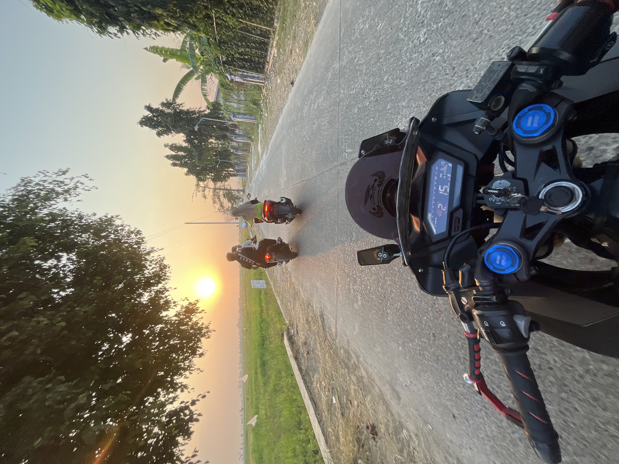 The image is a first-person perspective from a motorcycle in Pecangaan, Indonesia, during sunset. Two other motorcycles are visible ahead on a paved road. The motorcycle in the foreground displays a digital dashboard showing "2 15" with other indicators. The environment includes a green rice field to the left, bordered by a concrete curb, and trees and vegetation lining the right side of the road, with a fence and a building in the distance. The sun is setting, casting a warm glow over the scene.