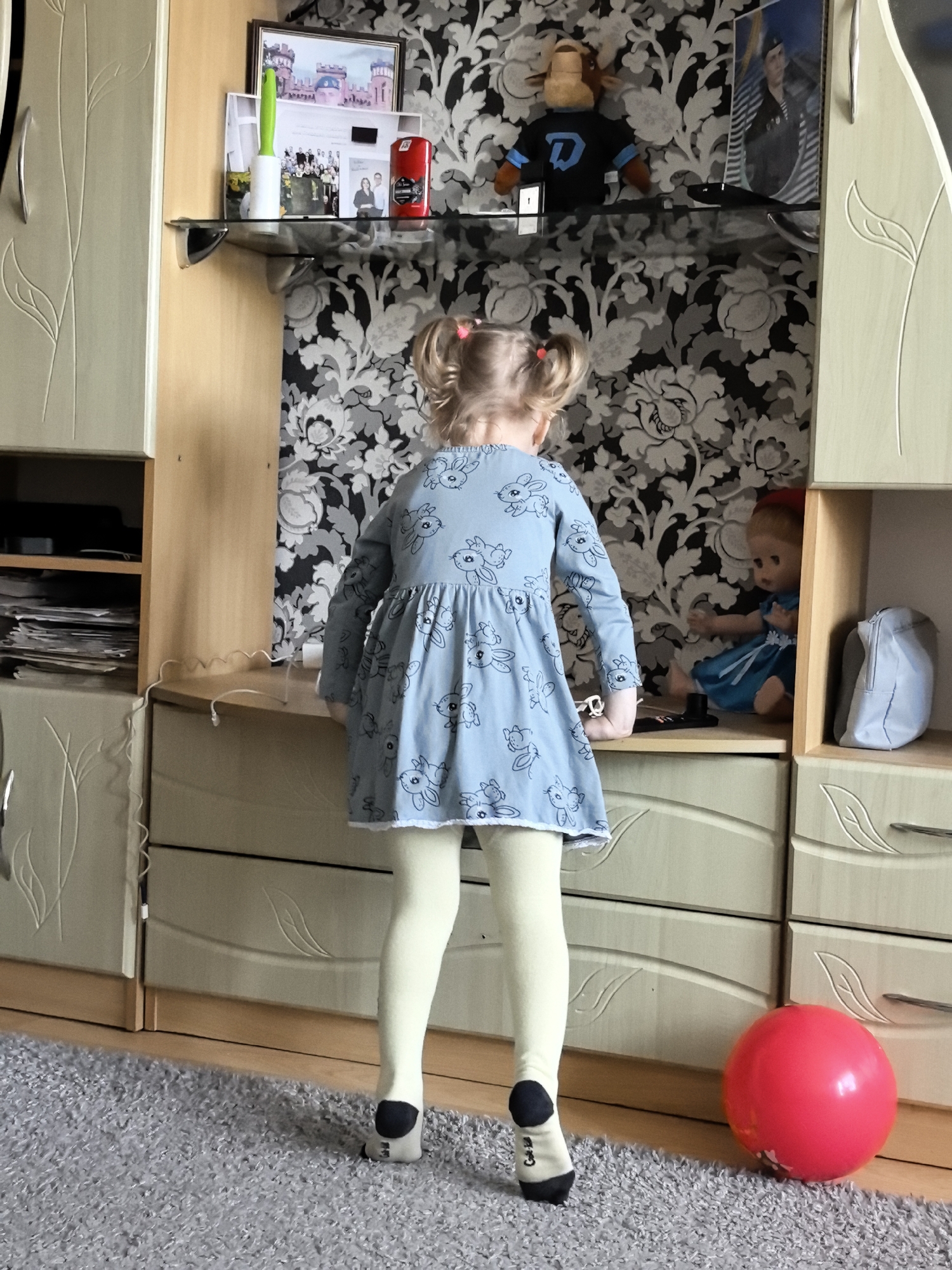 A full shot shows a young girl from the back, standing in front of a wall unit in an indoor setting. The girl is wearing a light blue dress with a pattern of cartoon rabbits and yellow leggings. Her hair is styled in two pigtails.
The wall unit is light beige with wooden accents. It contains shelves displaying various items, including framed photographs, a plush toy, a doll in a blue dress, and a light blue bag. On the lower part of the unit, there are drawers. A red balloon is visible on the gray carpet in front of the wall unit. The background features patterned wallpaper in black and white.
