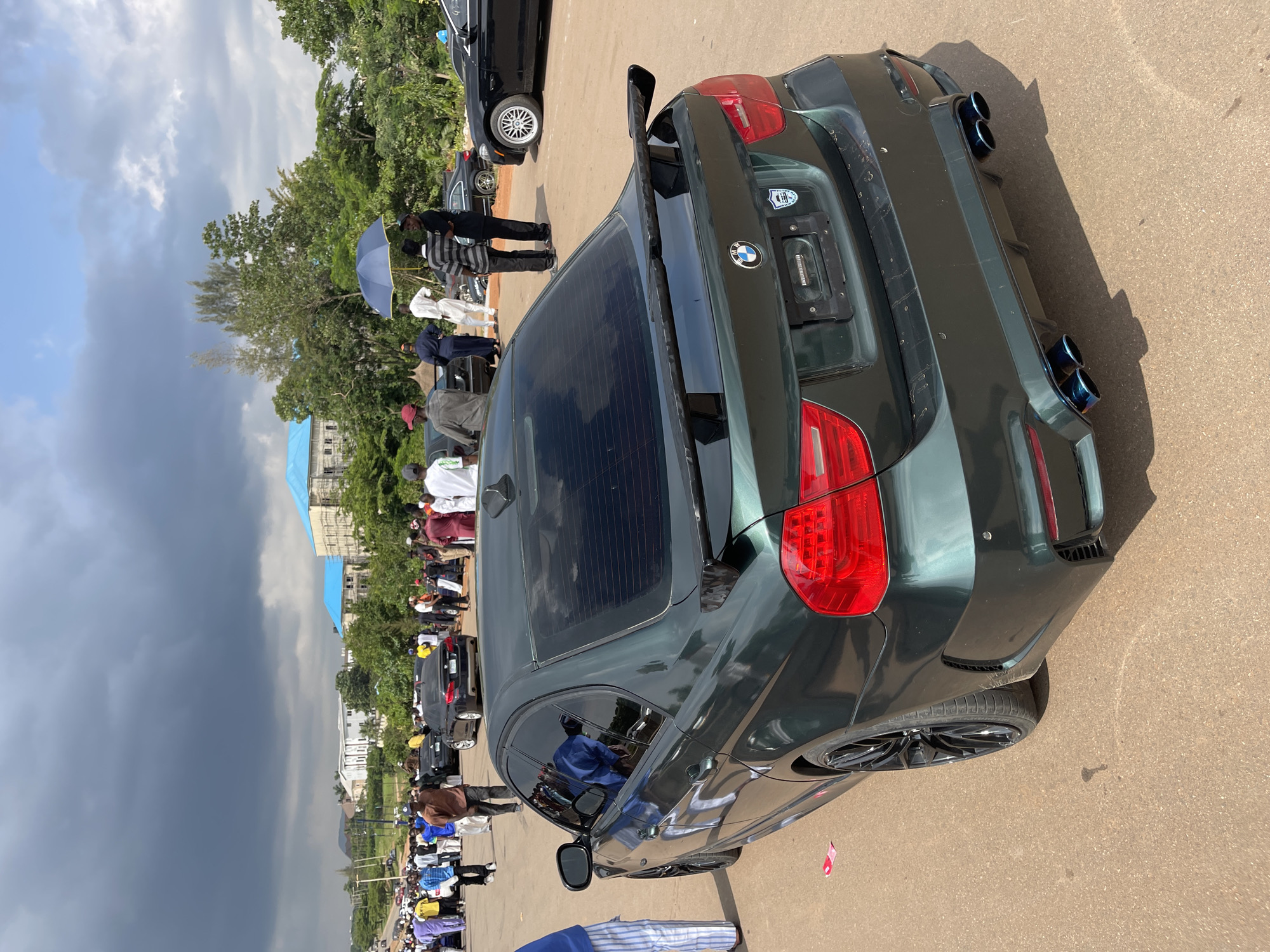 A dark green BMW sedan is parked on an asphalt road with a crowd of people in the background. The car has a spoiler and quad exhaust pipes. Some people in the background are holding umbrellas, and there are buildings with blue roofs visible in the distance. The sky is cloudy. The scene appears to be in a public area in Abuja, Nigeria, likely a gathering or event.