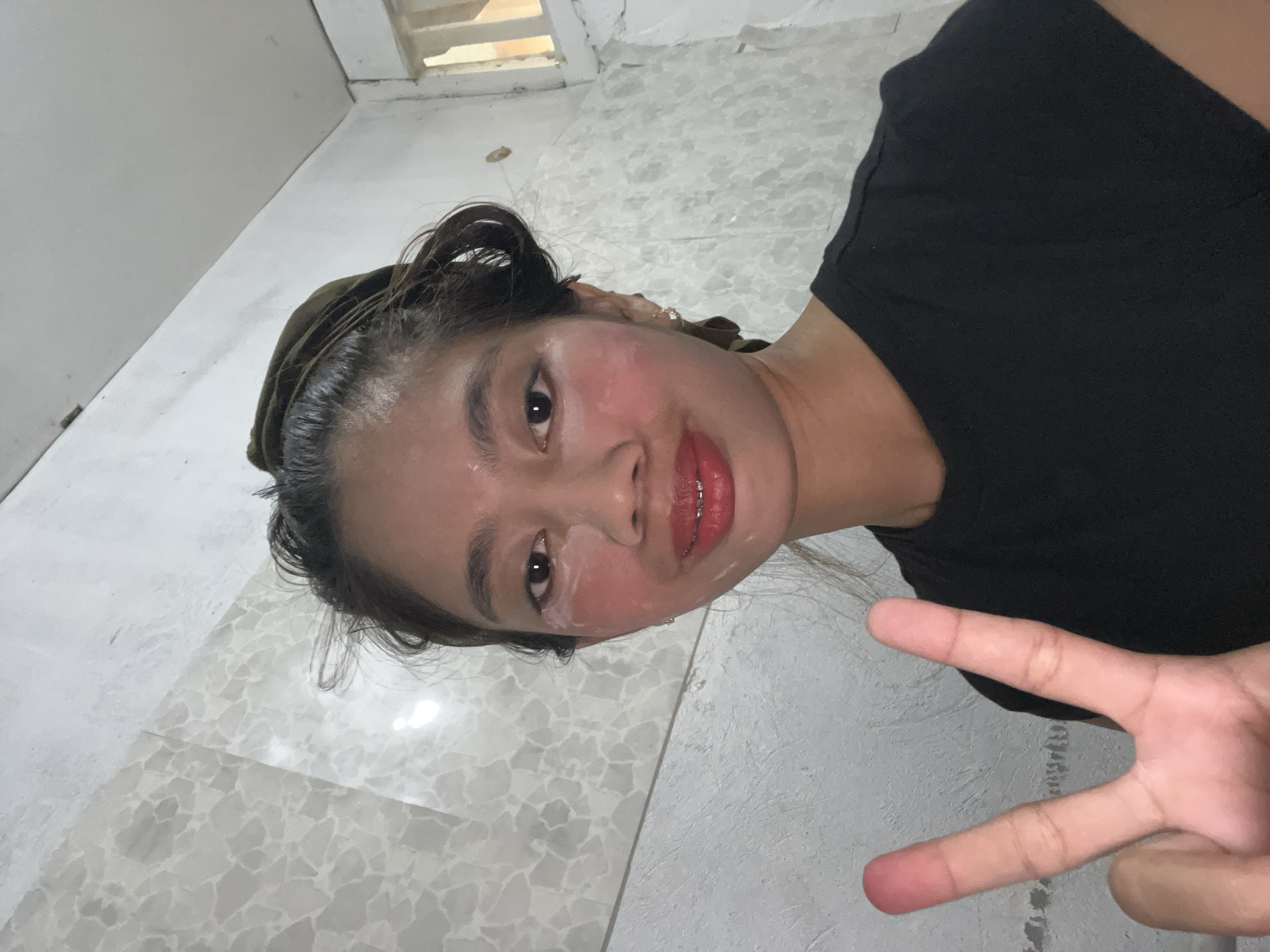 An indoor photograph, taken from a low angle, features a young woman posing, possibly in Davao City, Philippines. She has dark hair, pulled back with a dark green headband or scarf, and wears a black t-shirt. Her face is covered in a white, creamy substance, suggesting a skincare routine like a face mask, with her cheeks appearing flushed pink. She has bright red lipstick and visible dental braces on her upper teeth. Her left hand is raised in the foreground, displaying a peace sign.
The background shows a light-colored tiled floor with a mosaic or terrazzo-like pattern of irregular shapes, and a plain light grey/white tiled wall. A small opening or window with horizontal slats is visible near the top right corner. The lighting is soft and ambient. The woman's expression is neutral to slightly smiling, conveying a relaxed and casual demeanor. No text is visible.