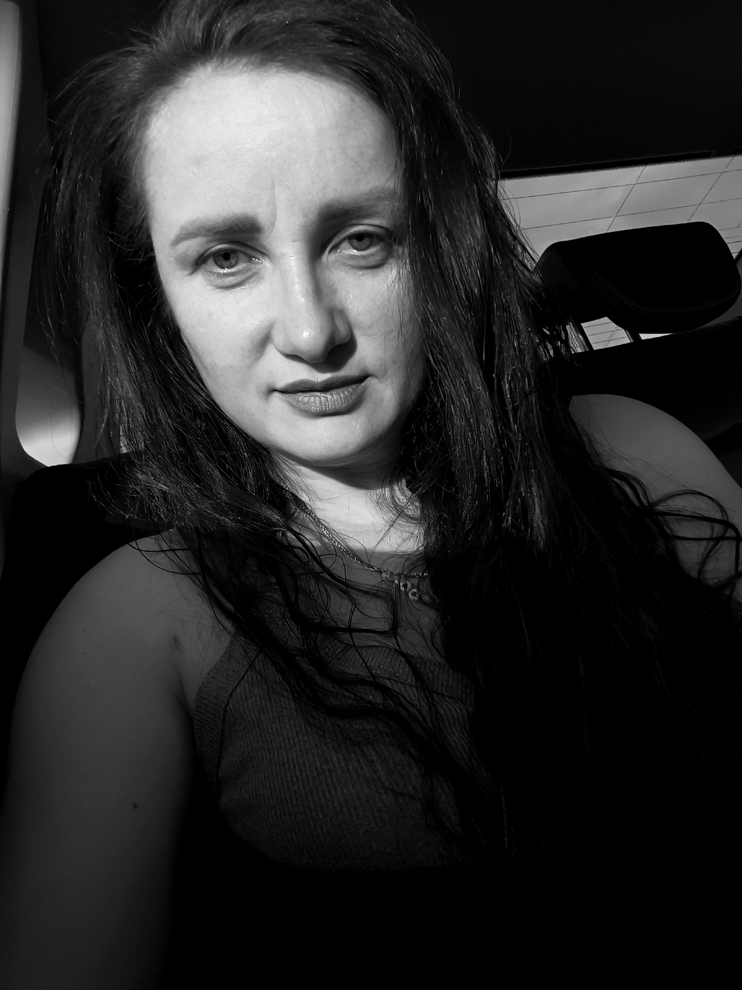 A black and white, medium shot, eye-level, selfie of a woman in a car at night. The woman has long, dark, wet hair that frames her face and falls over her shoulders. She is wearing a dark, ribbed tank top and a necklace. Her eyes are open and looking directly at the camera, with a neutral expression. Her lips are slightly parted. The background of the image is dark, with the back of a car headrest visible to the right. The overall lighting is dim, with shadows cast across the woman's face and body. The car appears to be stationary, possibly parked in a dark area of Lelystad, Netherlands.