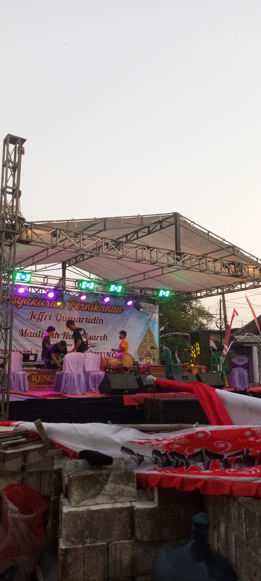 The image depicts an outdoor stage setup for a wedding reception or similar event in Paji, Indonesia. The stage is constructed of metal scaffolding and covered with a white canopy. Colorful lights, green and purple, illuminate the stage from above. A large banner with text, including "SYUKURAN PERNIKAHAN" (Wedding Thanksgiving) and names, is displayed behind the performers.
On the stage, three young men are present. One, wearing an orange shirt, is playing a traditional drum (gamelan or similar). Another man in a black t-shirt is standing and appearing to adjust something on the stage. A third man, in a dark shirt, is crouched down, also seemingly working on equipment. There are also two chairs draped in purple fabric, and various sound equipment, including speakers.
In front of the stage, there is a low wall made of stacked bricks, some of which appear weathered. A red and white fabric with a swirling pattern is draped over some of this wall, suggesting it is part of the event decorations or a makeshift barrier. The background shows trees and a building with a red and white flag, indicating a festive atmosphere. The sky is overcast with a pale, washed-out color. The overall scene suggests a community gathering or celebration taking place outdoors in the late afternoon or early evening.