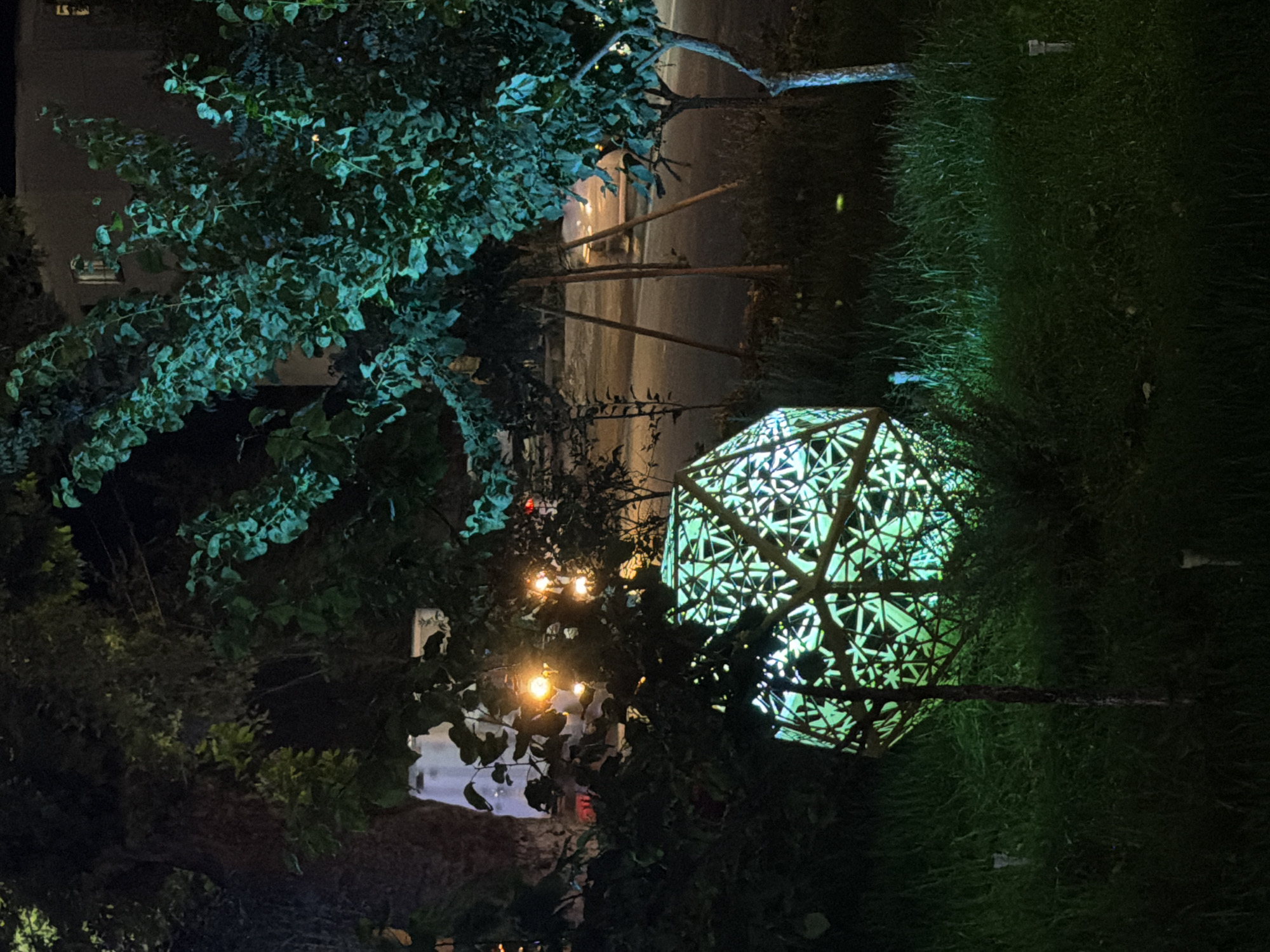In the city of Chirchik, Uzbekistan, at night, a large, faceted, geometric lantern glows with a vibrant green light. The lantern, made of a metal lattice, is situated on a grassy embankment, its intricate design casting patterned shadows. It is surrounded by lush green grass and foliage. In the background, trees with leaves illuminated by what appears to be teal lighting create an ethereal atmosphere. Beyond the trees, a street is visible with some streetlights casting warm yellow and orange glows. A building with visible windows can be seen in the distance. The scene is illuminated by artificial lighting, suggesting a public or park area.