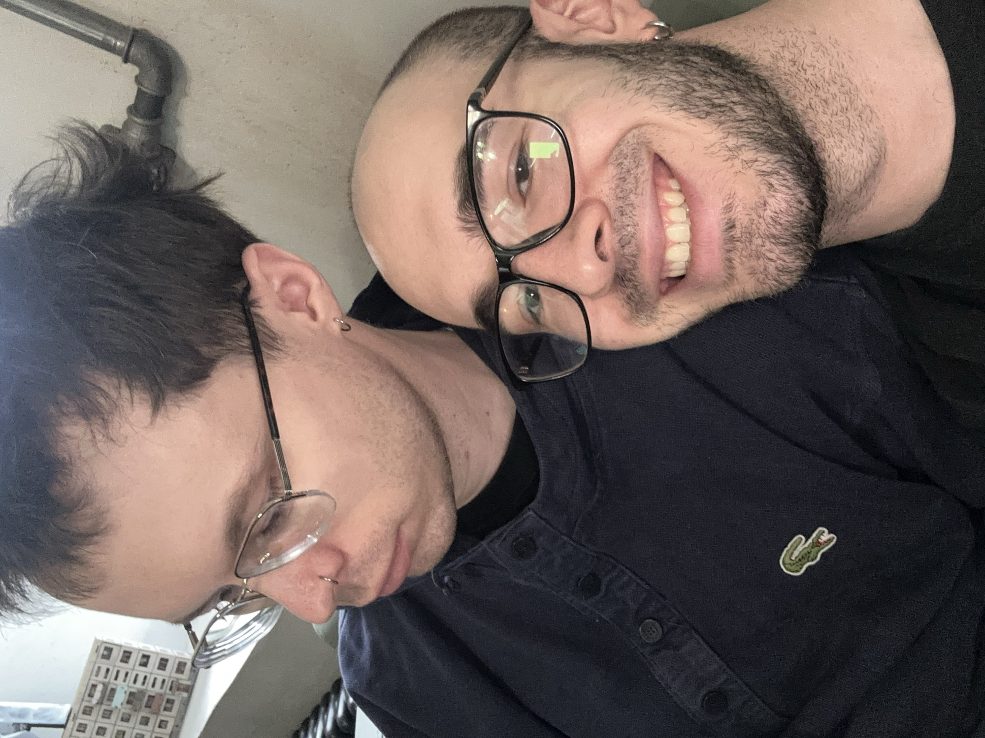 The image depicts two men in close proximity. One man is partially bald with a beard and glasses, smiling broadly. He is wearing a dark shirt with a Lacoste logo. The other man, with dark hair and glasses, appears to be looking down. He also has a piercing on his nose. They are posed closely together. A metal pipe is visible near the top left of the frame. A box of what seems to be film slides or other small objects is visible in the lower left corner. There are no features in the image to indicate that this was taken in Moscow, Russia.