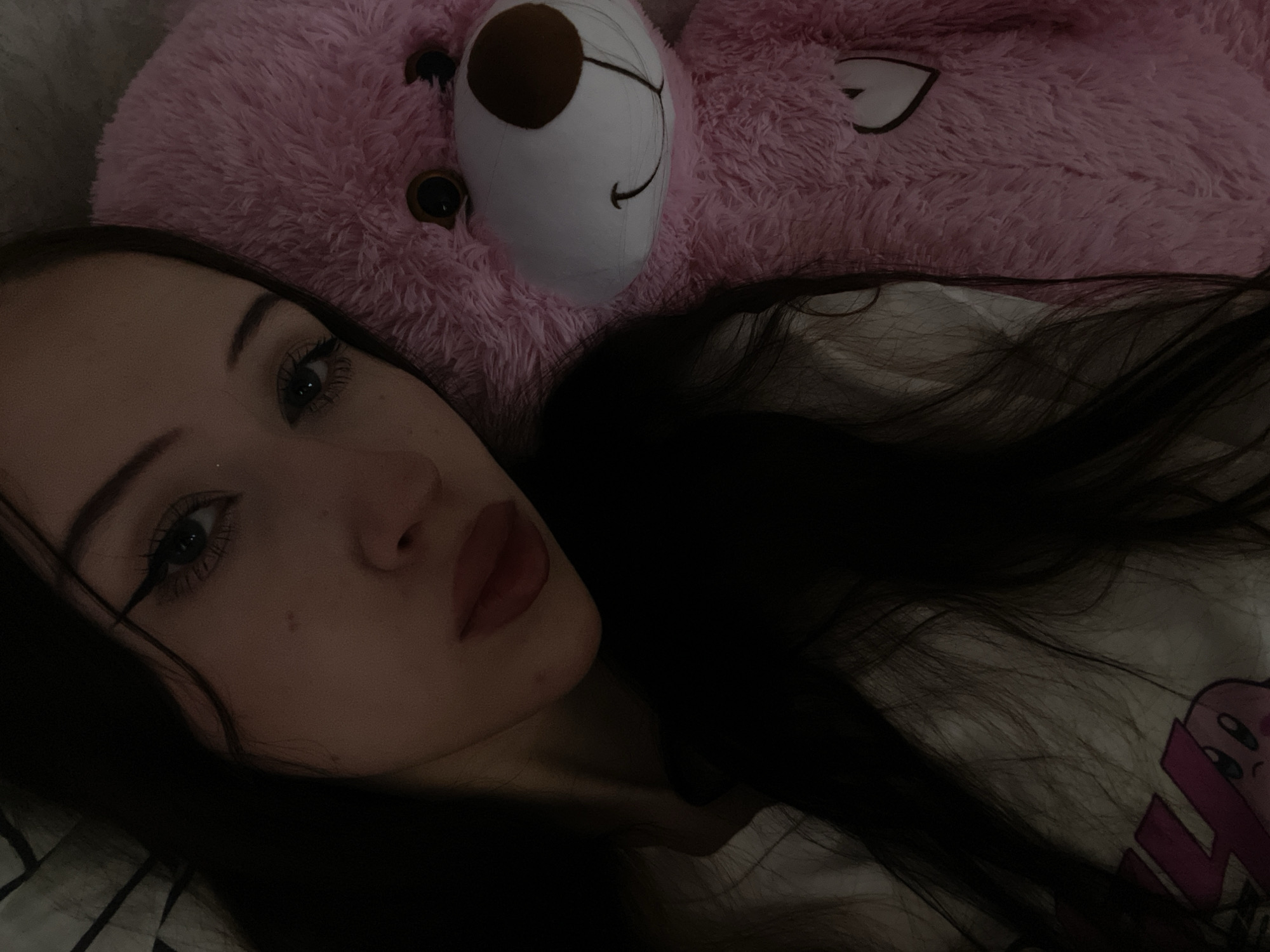 A close-up selfie taken indoors, featuring a young woman with long dark hair and makeup, resting her head next to a large, fuzzy pink teddy bear. The woman's eye, nose, and lips are visible. She is wearing a white t-shirt with a character graphic. The teddy bear is positioned to her right and fills a significant portion of the frame, displaying a stitched heart on its chest. The lighting suggests it is nighttime or a dimly lit room. No specific location in Votkinsk, Russia can be determined from the image.