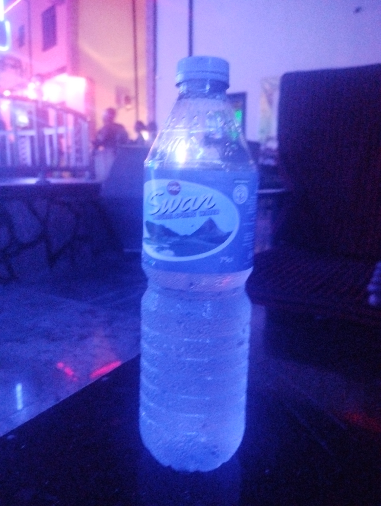 The image shows a bottle of Swan Spring Water, 75cl, sitting on a dark surface. Condensation is visible on the bottle. In the background, there's a blurred view of what appears to be a bar or lounge with people present. The lighting is predominantly blue. The image does not provide sufficient information to confirm a location in the city of Tumfure, Nigeria.