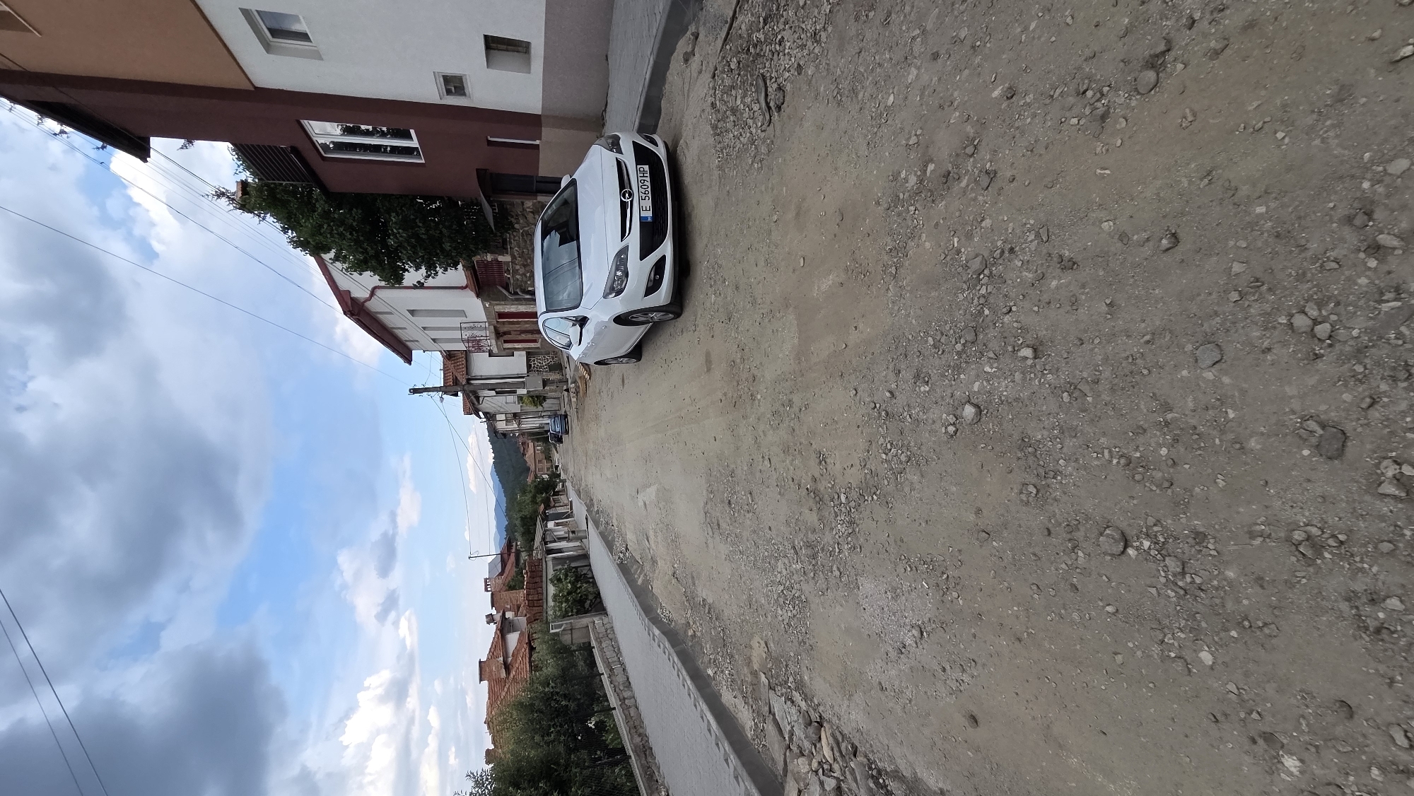 The image depicts a street scene in the city of Bania, Bulgaria, characterized by ongoing road construction. The road surface is unpaved, composed of dirt and gravel, with scattered stones and debris. A white Opel Astra automobile, license plate E 5609 HP, is parked on the right side of the street. The left side of the street features a newly constructed sidewalk with gray paving stones and a curb. Adjacent to the sidewalk are residential buildings with terracotta tile roofs. The sky is partly cloudy, with a mix of gray and white clouds against a blue background. In the distance, rolling hills and mountains are visible. The overall impression is one of a rural or semi-urban setting undergoing infrastructure development.