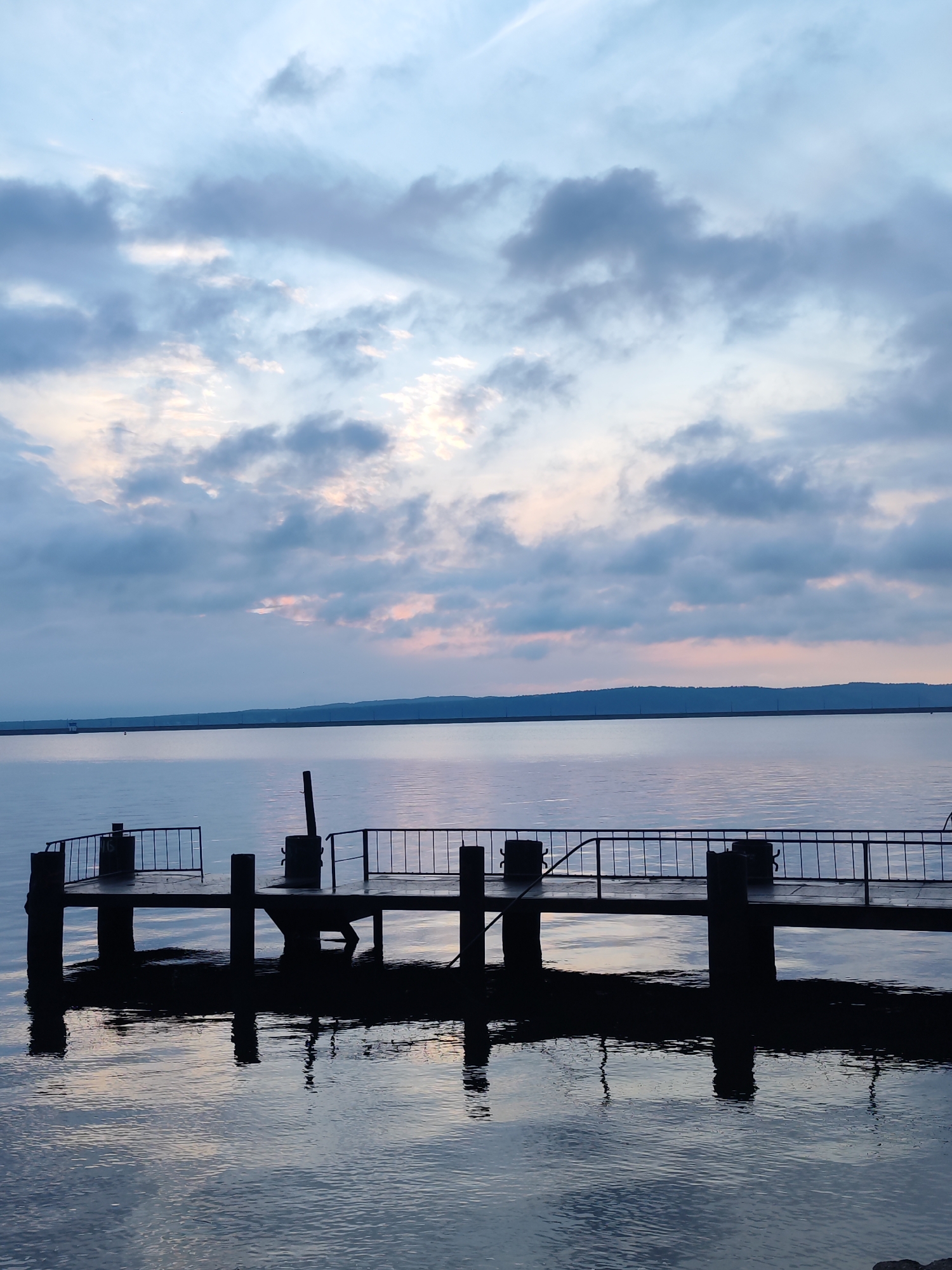 The image depicts a pier extending into a body of water, likely a lake or reservoir, under a cloudy sky at dusk or dawn. The pier is constructed of dark-colored material, with metal railings along portions of its sides. Vertical posts support the pier's structure, and these posts are reflected in the water below. The water's surface is calm, displaying subtle ripples and reflections of the sky and pier. In the background, a distant landmass is visible, with a subtle change in elevation suggesting hills or low mountains. The sky is filled with clouds ranging in color from gray to bluish, with hints of pink or orange near the horizon. This location is in the city of Chaikovsky, Russia. There are no people visible in the scene.