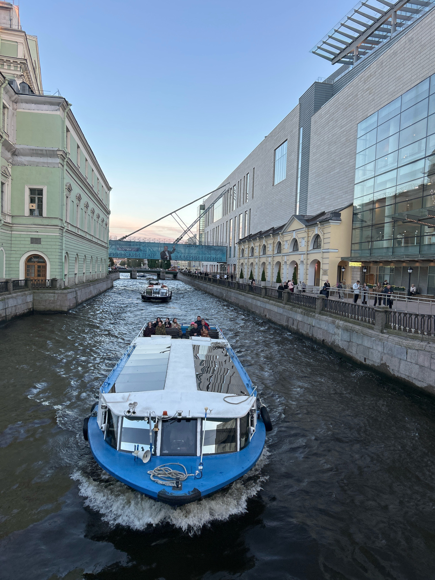 A high-angle, medium shot captures a blue and white tour boat navigating a canal in an urban setting under a clear, pale sky. The boat, with a white cabin and blue hull, is in the foreground, leaving a white wake as it moves forward. The boat is carrying passengers who are visible on its upper deck.
In the middle ground, two smaller boats are also on the canal, one with passengers, the other appearing to be a service boat. A bridge spans the canal in the distance, with a large banner advertising the Mariinsky Theatre.
On the left bank of the canal is a pale green building with a classical facade and arched windows. On the right bank, a modern building with large glass windows and a stone exterior rises up, adjacent to a row of yellow buildings with arched doorways. People are walking along the embankment on the right side of the canal, some leaning on a railing. The canal walls are made of stone. The overall scene suggests a day of light activity on the water in a city.