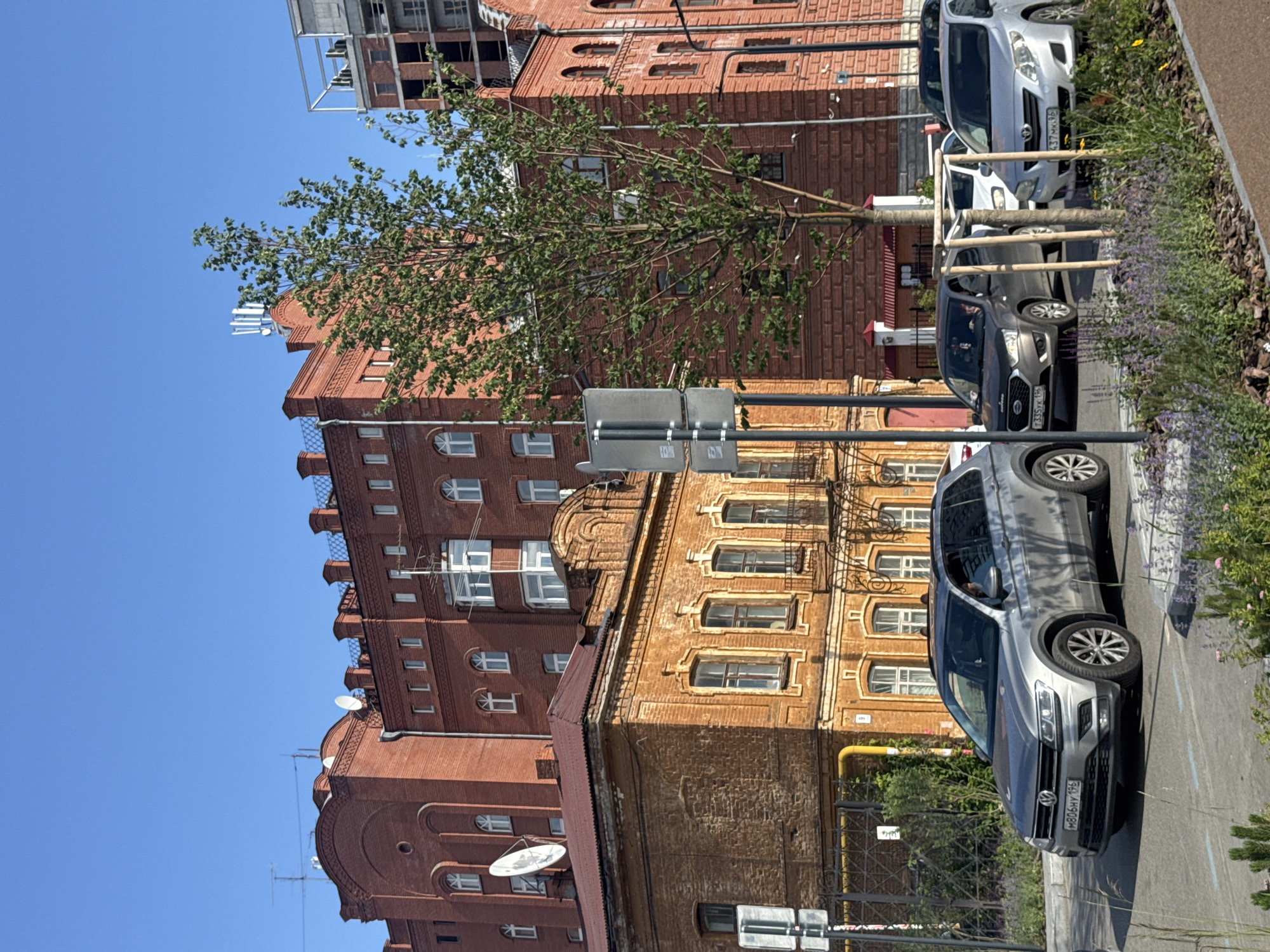 The image displays a street scene in Yekaterinburg, Russia, on a clear, sunny day. The focal points are several buildings and parked cars. Dominating the scene are two brick buildings; the one on the left is a darker red, and the one on the right is a lighter, more golden hue, showcasing intricate architectural details around its windows and cornices. A young tree with green leaves is positioned in the foreground, obscuring part of the buildings.
Several cars are parked along the street. A silver SUV is prominently featured in the mid-ground, with its front facing towards the viewer. To its left, a dark grey car is visible, and further back, a light silver car is parked. The license plate on the grey car is partially visible and reads "M806HY 196". A signpost with two rectangular signs is also present, mounted on poles.
The architecture suggests an urban environment, possibly a historic district, with traditional brickwork and decorative elements on the buildings. The clear blue sky and bright sunlight indicate it is daytime. The overall impression is that of a typical urban street scene on a pleasant day.