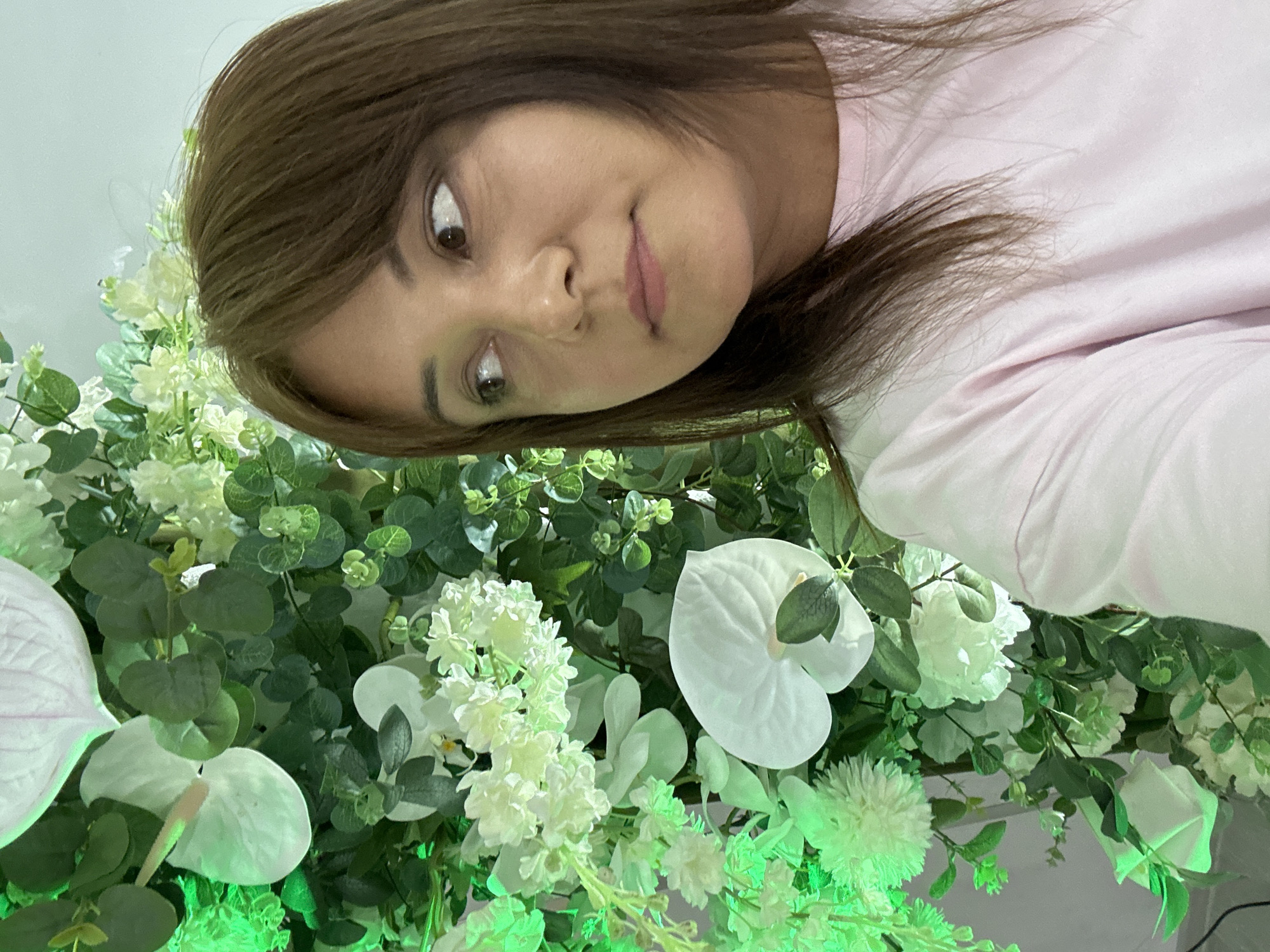 A woman with brown hair and a pink shirt is positioned in front of a flower arrangement. The arrangement includes white Anthurium flowers, white hydrangeas, eucalyptus leaves, and other greenery. The lighting on the flowers appears to be tinged with green. The woman looks slightly downwards with a curious expression. The location appears to be an indoor setting. The city is Unknown, Unknown.