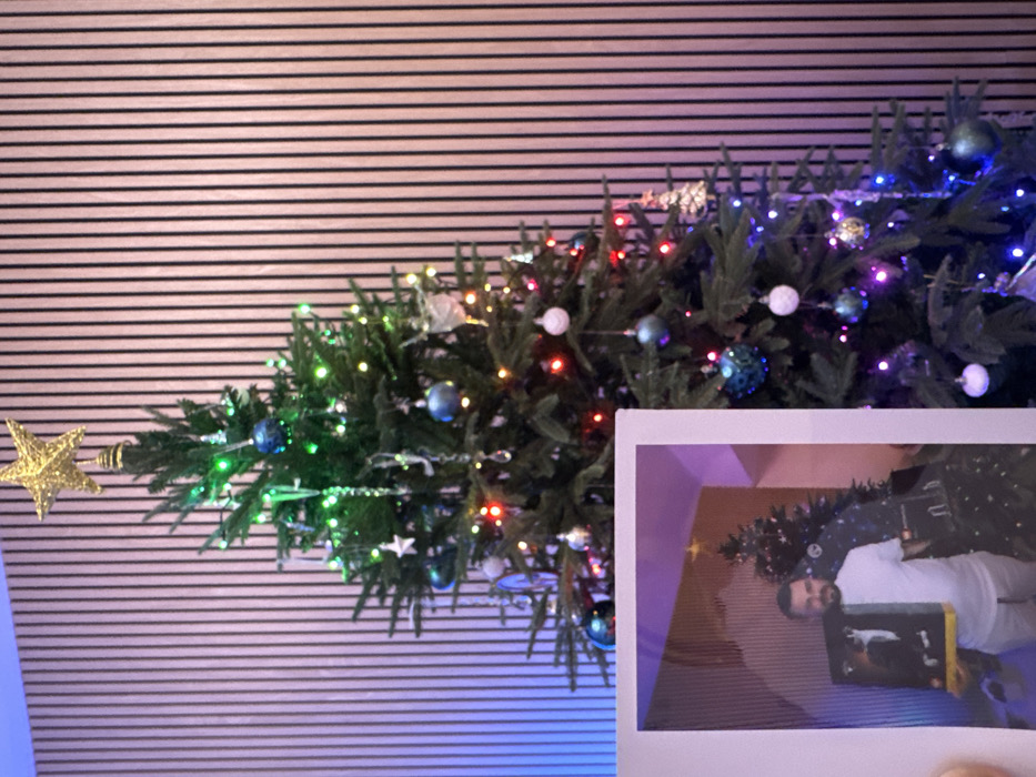 An indoor scene in Dubai, United Arab Emirates, features a decorated Christmas tree positioned against a light-colored, vertical slatted wall. The artificial evergreen tree is adorned with a golden, glittery five-pointed star topper, multicolored string lights (red, green, blue, purple), and various ornaments including blue, white, and teal spherical baubles, white stars, and clear icicle-shaped drops. Ambient blue light illuminates the left portion of the slatted wall. In the lower left foreground, a white-bordered instant photo print is held, partially obscuring the Christmas tree. The print displays an adult male with short dark hair and a beard, wearing a white short-sleeved t-shirt and light trousers. He holds a dark rectangular object with a yellow base, featuring a white figure, in front of him. Behind the man in the print, another decorated Christmas tree is visible against a vertically slatted wall, illuminated by purple and orange ambient light, indicating a similar interior environment.