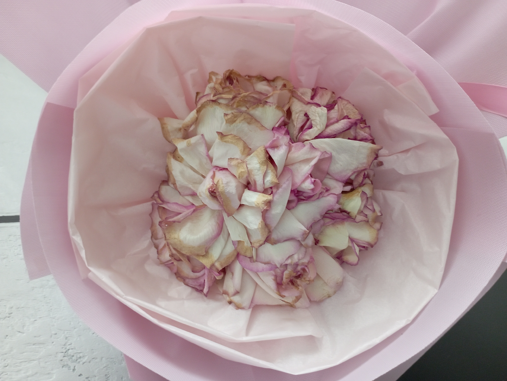 A close-up, top-down shot shows a wilting bouquet of pink and white roses, their petals dried and curled with brown edges. The roses are nestled within a pink paper wrapping that has a slightly textured outer layer and a smoother, tissue-like inner layer. The bouquet is set against a white, textured background, possibly a wall. There are no people or animals visible in the image.