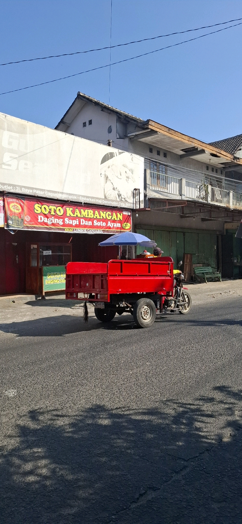 In Desa Sawahan, Indonesia, an outdoor scene unfolds under a clear blue sky. A bright red, three-wheeled truck with a trailer is parked on a paved road, its trailer containing a blue umbrella and some items. The truck is positioned in front of a building with a large banner advertising "SOTO KAMBANGAN" and "DAGING SAPI DAN SOTO AYAM." The building appears to be a shop or restaurant, with green shutters visible on the lower level and balconies on the upper level. Shadows of trees are cast on the road, suggesting the presence of foliage nearby. The overall impression is of a commercial area in a town or village.