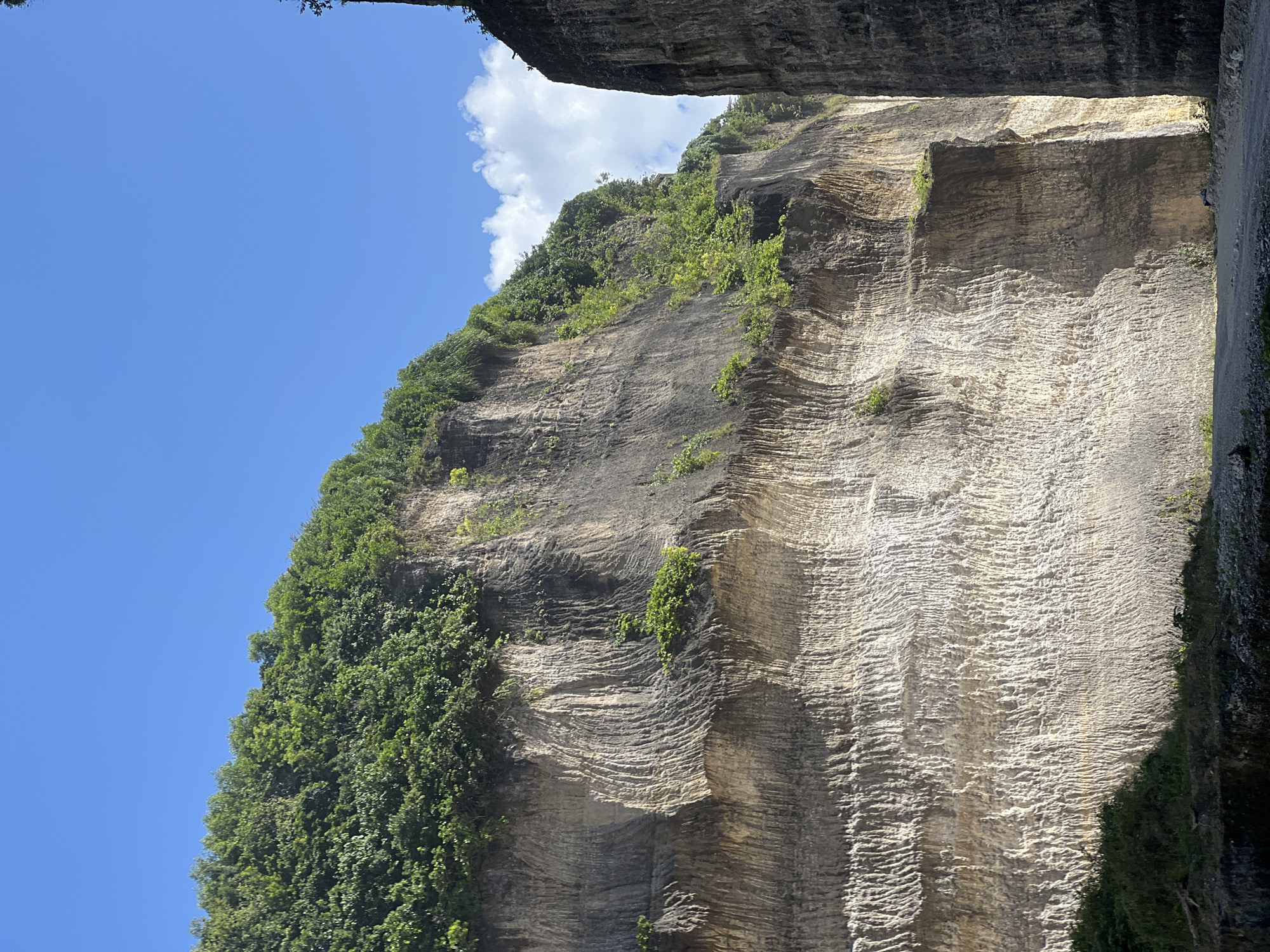 The image depicts a limestone cliff face in Kuta Selatan, Indonesia. The cliff is layered and textured, with vegetation clinging to sections of the rock face. The sky is blue with a few white clouds visible. A portion of the cliff overhangs in the top of the frame.