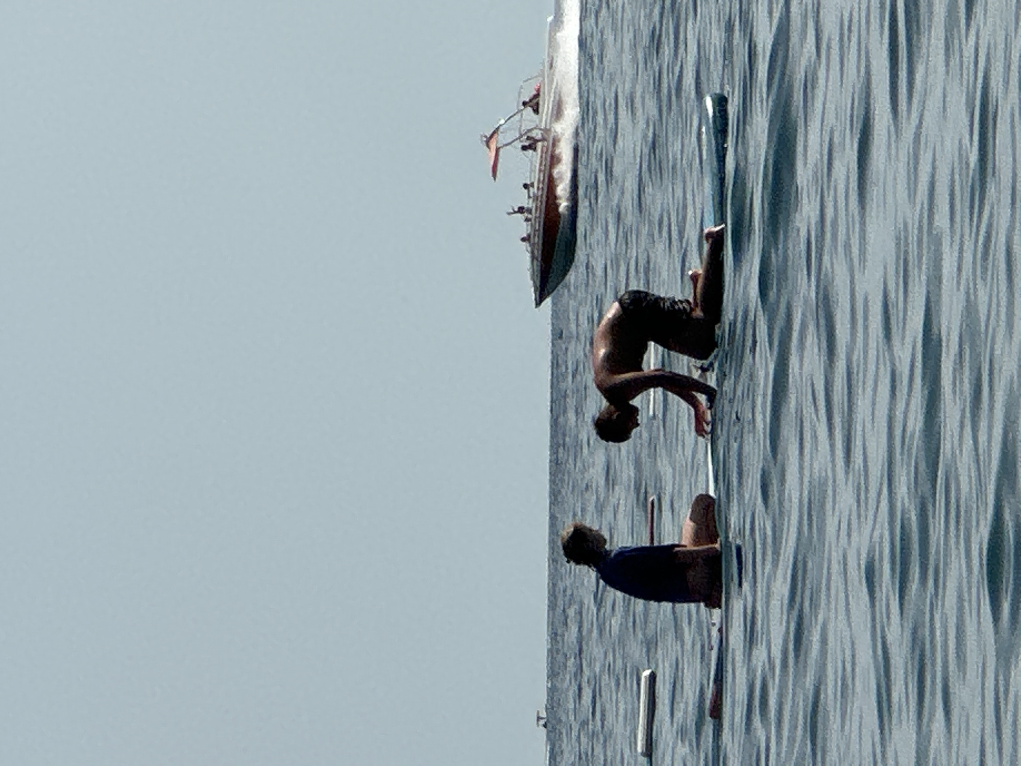 Two people are on paddleboards in the water. One person is kneeling and facing away from the camera, with their arms outstretched. The other person is sitting down and facing the kneeling person. In the background, a boat with several people on it is moving through the water. The sky is clear and blue. The scene appears to be in an outdoor setting, likely on a lake or the sea. The location is specified as Boreti, Montenegro.