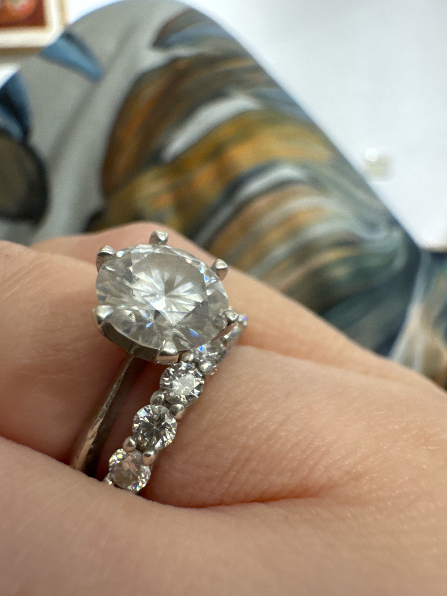 A close-up, slightly angled shot shows a person's hand wearing a white gold engagement ring and a diamond-encrusted wedding band. The engagement ring features a large, round, brilliant-cut diamond set in a four-prong setting. The wedding band is a delicate band with several small, round, brilliant-cut diamonds. The background is blurred, showing a swirled pattern of gray, blue, and tan colors, possibly fabric or a painted surface. The lighting is bright, highlighting the sparkle of the diamonds. The location of this image in Tomilino, Russia is not discernible from the visual information provided.
