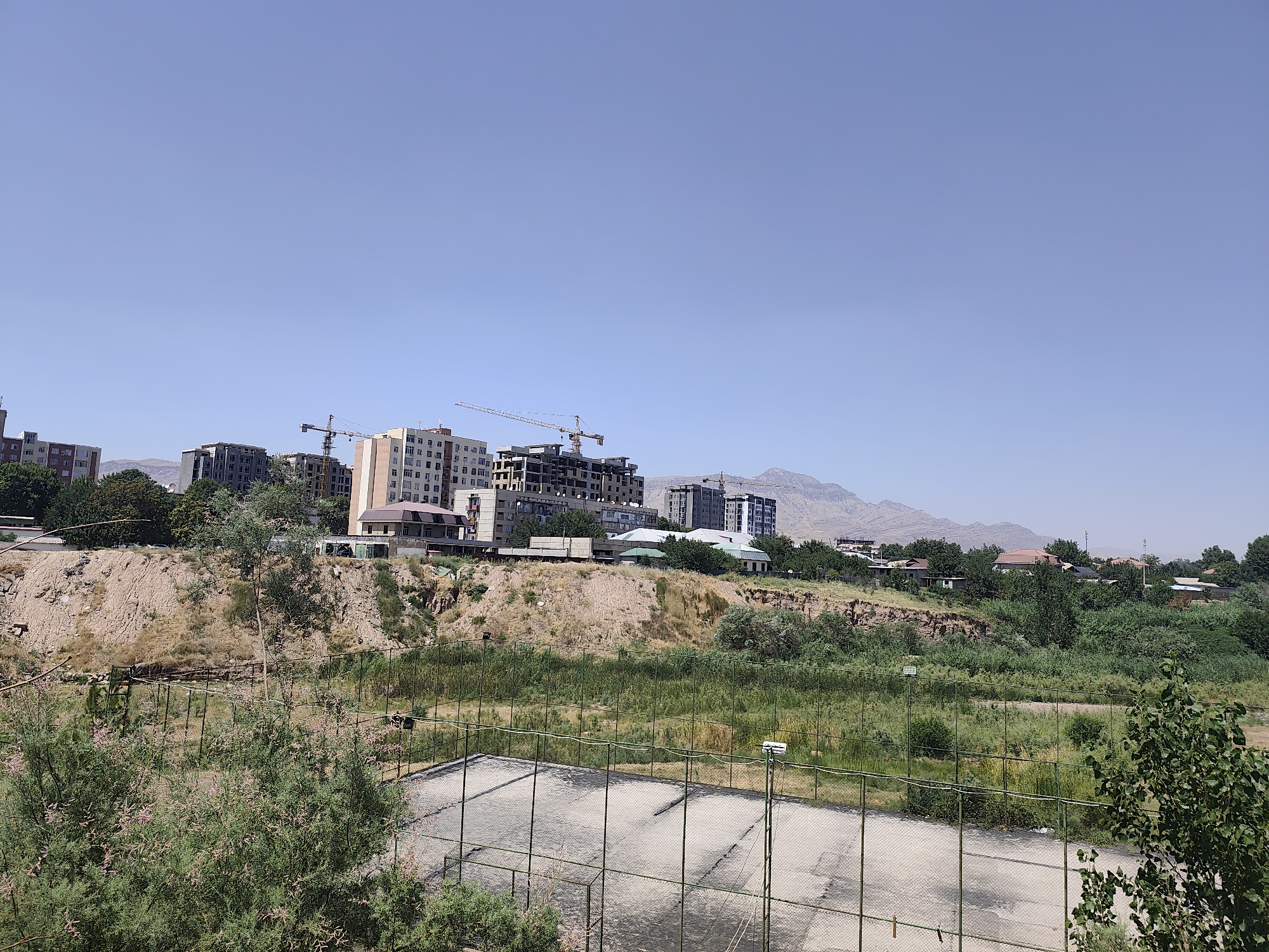 The image depicts a daytime scene in the city of Yovon, Tajikistan, showcasing a blend of urban development and natural landscape. In the foreground, a fenced-off, weathered outdoor court with a concrete surface is visible, surrounded by green foliage. Beyond this area, a steep, dry, earthen embankment rises, dotted with sparse vegetation.
Dominating the midground and background is a developing urban skyline. Several multi-story buildings, some appearing completed and others under construction with visible cranes, are situated on the elevated terrain. The architectural style suggests modern apartment complexes. A further expanse of greenery, including trees and shrubs, separates the embankment from the more distant residential areas and the mountainous backdrop. The sky is clear and blue, indicating fair weather.
