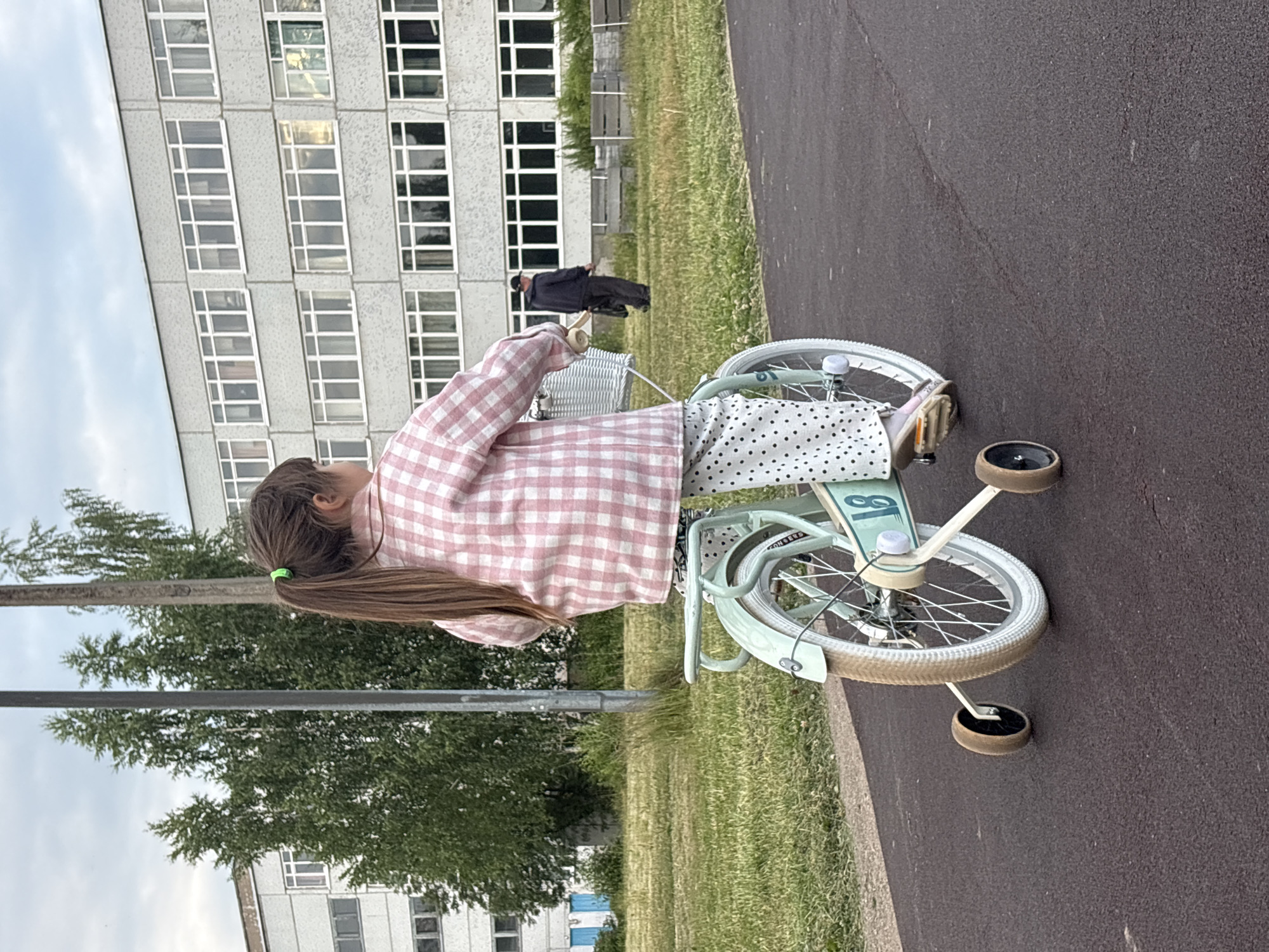 A young girl with long brown hair tied in a ponytail with a green elastic band is seen from behind, standing next to a mint green bicycle with training wheels. She is wearing a pink and white checkered shirt and a white skirt with black polka dots. The bicycle has a white basket in the front. In the background, a man in a dark jacket and cap is walking. To the left of the girl and the man, there is a large, modern, multi-story building with many windows. The building appears to be made of concrete. Further in the background, there are trees and a cloudy sky. The scene appears to be outdoors, on a paved path next to a grassy area. The overall impression is that of a casual, possibly late afternoon or early evening setting. The image might be taken in Chaikovsky, Russia, given the location context. There is some text visible on the bicycle, but it is not clearly legible.