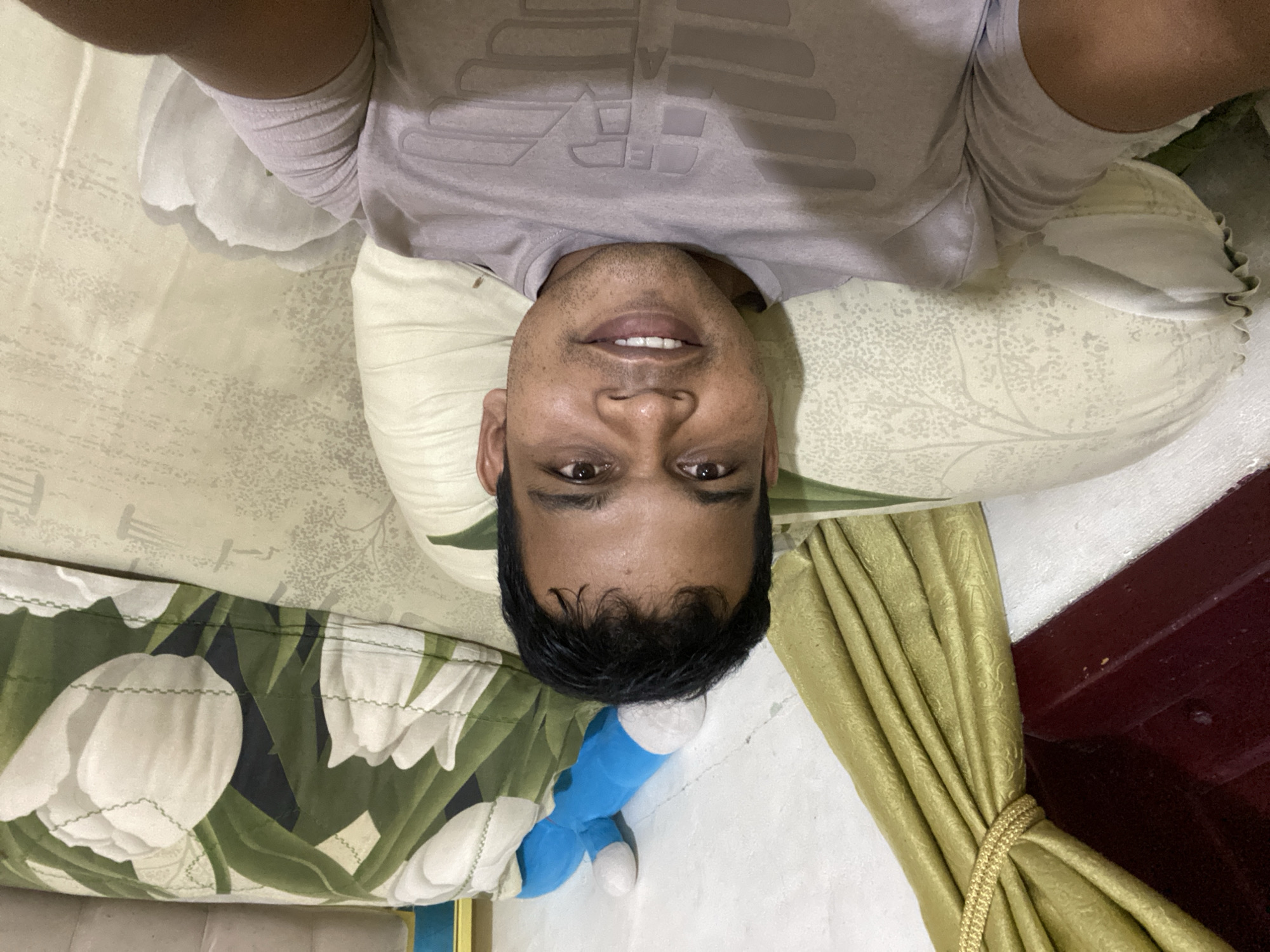 A medium-shot, selfie-style photo shows a dark-skinned man with short, dark hair and a light-colored t-shirt with a logo on the chest. He is lying down, with his head resting on a light-colored pillow. Behind him, a pillow with a floral pattern of white tulips and green leaves is visible. To the left, a gold curtain is partially drawn, revealing a dark red surface. A blue stuffed toy is propped up against a white wall in the background. The man is smiling and looking at the camera. The overall setting appears to be a bedroom.
