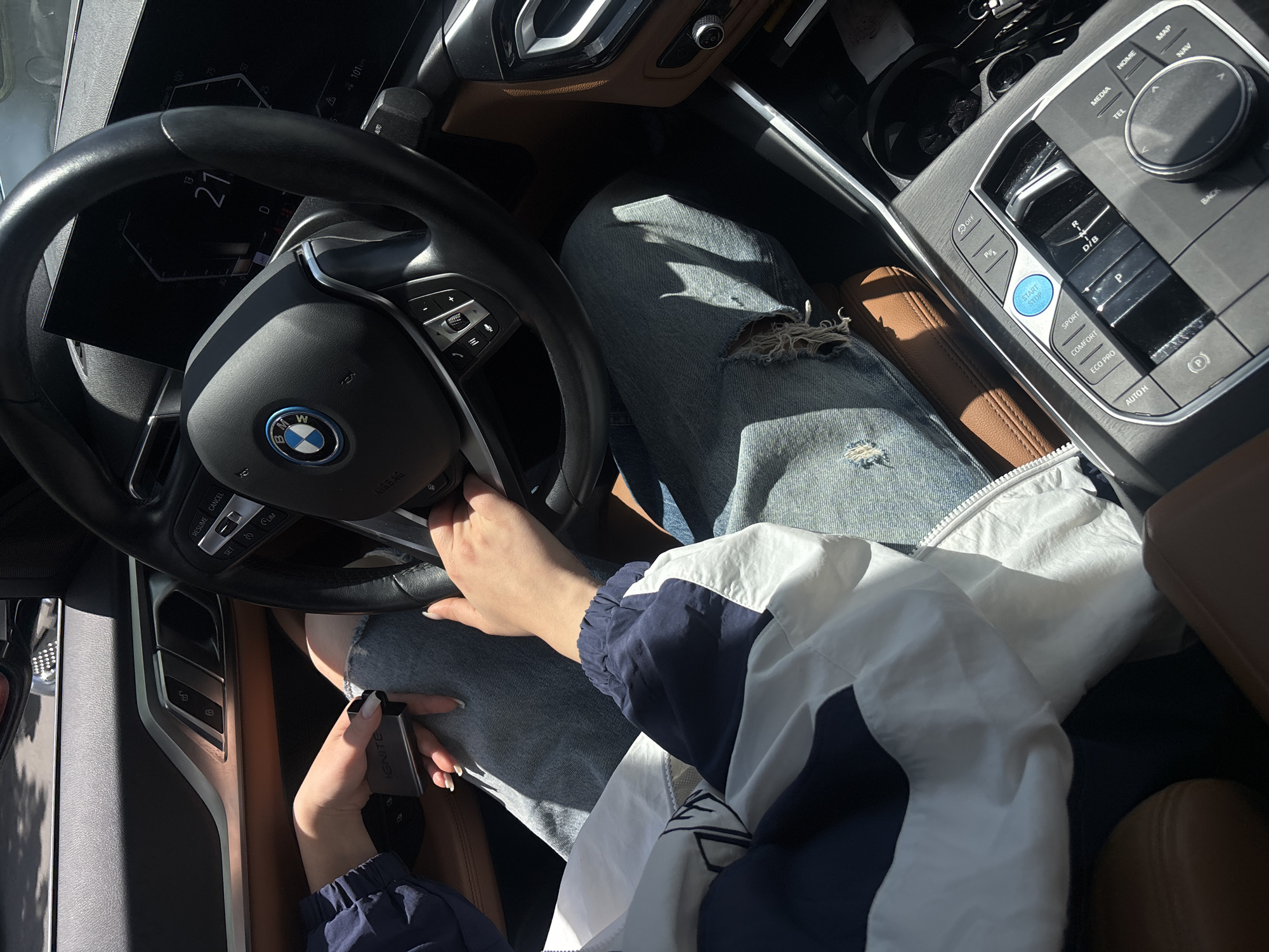 The image shows the interior of a BMW car. The driver is holding an "IGNITE" device and has their hand on the steering wheel. The car's dashboard indicates a speed of 27 km/h and a remaining power of 101 km. The center console features various control buttons, including drive mode selectors (SPORT, COMFORT, ECO PRO) and infotainment controls. The driver is wearing blue jeans with a rip and a white and blue jacket. The environment appears to be sunny. There is no information about Yerevan, Armenia.