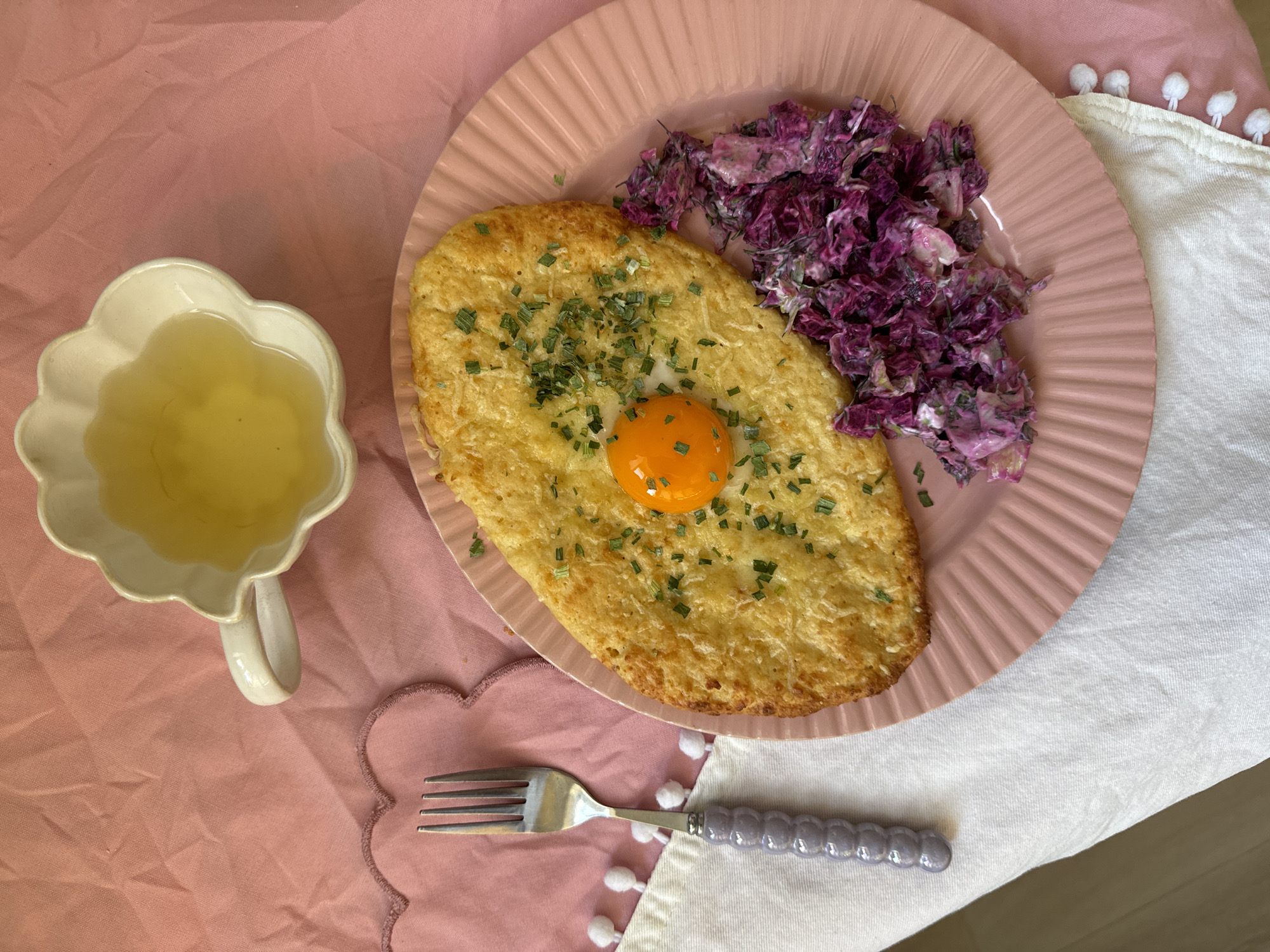 A high-angle shot depicts a meal arranged on a pink tablecloth, consisting of a pink plate with a fried egg centered on what appears to be a potato pancake or hash brown, sprinkled with chives. A serving of red cabbage slaw is to the right of the potato cake. A small white cup with a flower petal shape contains a clear liquid. A silver fork with a decorative grey handle lies below the plate. Part of a white napkin with pom-pom edging is visible to the right of the fork and plate. The background is a mix of pink and white fabrics. The location cannot be identified as Krasnoyarsk, Russia from the given image.