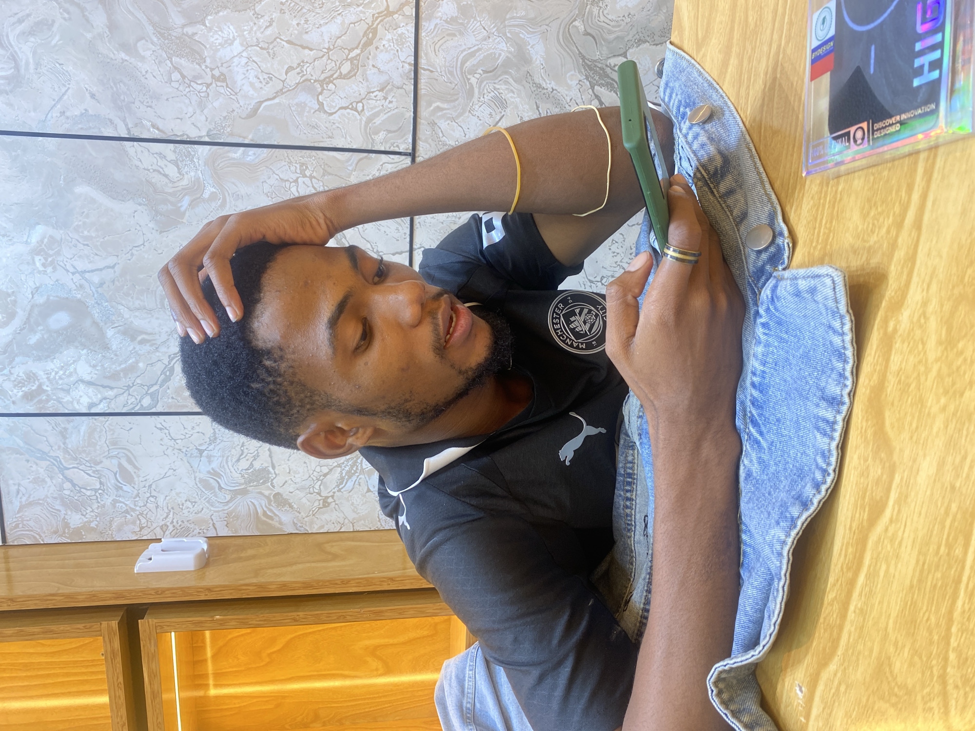A medium shot shows a man with dark hair, a beard, and a gold ring on his finger, wearing a black t-shirt with the Manchester City logo on the left side of his chest, looking at a green smartphone he is holding in his right hand. His left hand is raised to the top of his head. He is sitting at a table covered with a blue denim jacket. A package is also on the table near the right side of the image. The background consists of a beige marbled wall, and a wooden cabinet. The location is likely a cafe or restaurant in Dar es-Salaam, Tanzania.