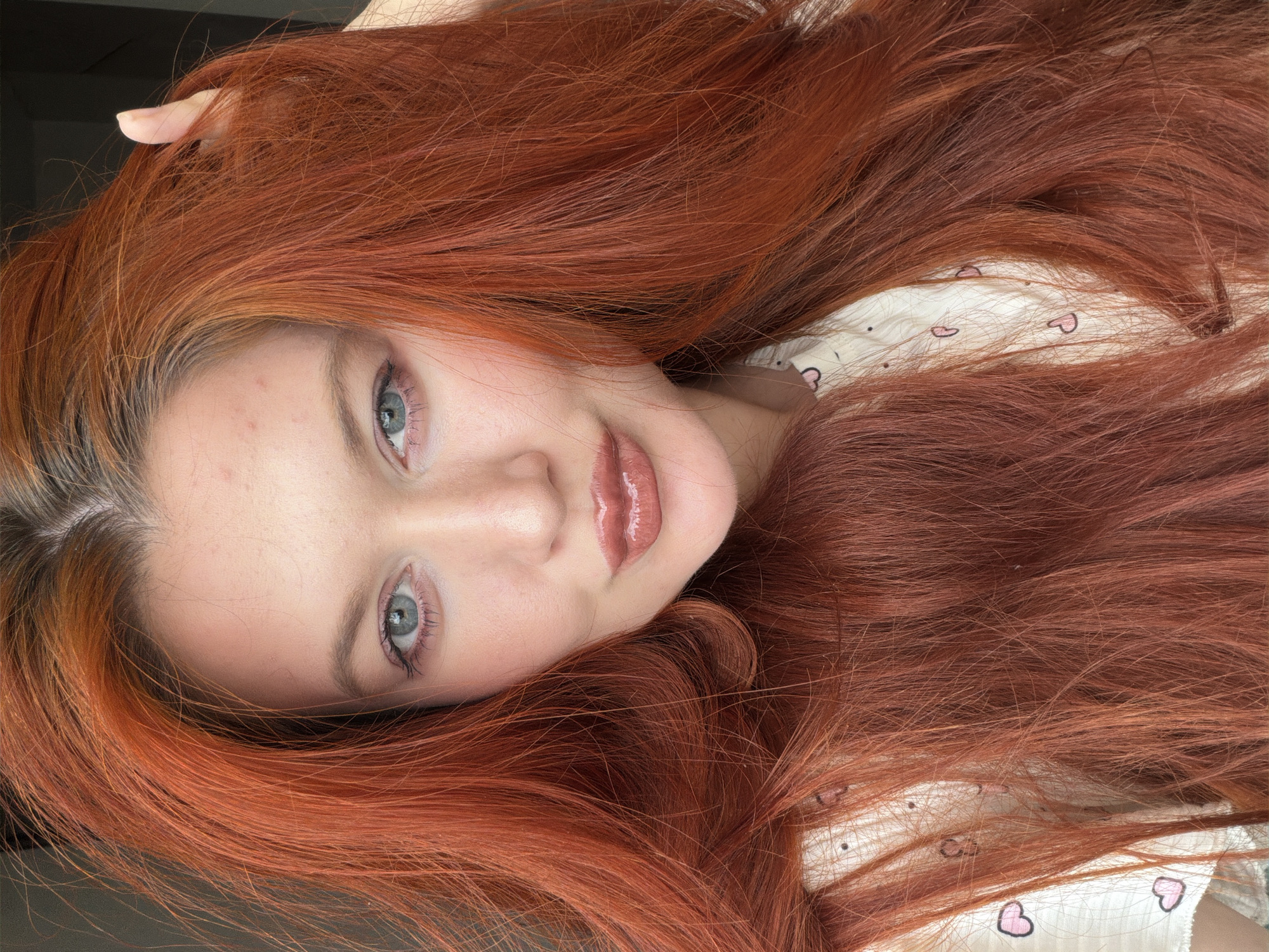 The image is a close-up, front-facing portrait of a woman with vibrant, long, wavy, red hair. Her hair is the dominant feature, framing her face and falling around her shoulders. She has striking blue eyes, light skin with a rosy complexion, and full lips with a glossy, neutral-toned lipstick. Her eyebrows are well-defined and arched. She is wearing a white top with a pattern of small pink hearts. Her left hand is visible, gently touching her hair near her temple. The background is dark and out of focus, with no discernible environmental details that would suggest a specific location in Krasnodar, Russia, or any other city. The lighting appears to be soft and even, highlighting the texture and color of her hair and facial features.