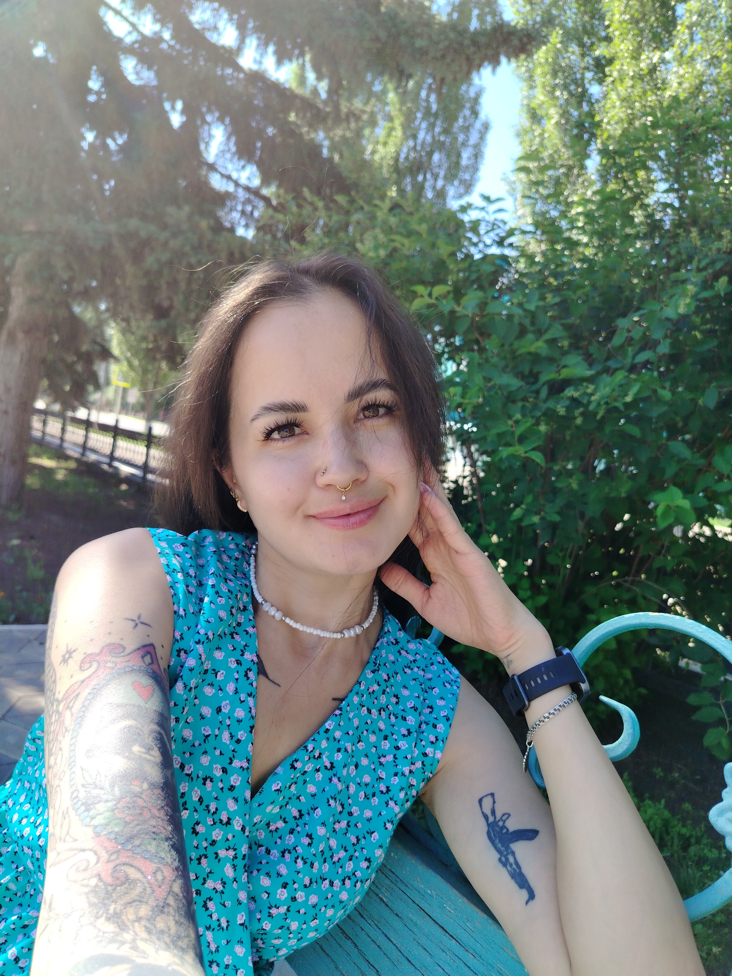 A young woman with dark hair, brown eyes, and fair skin is sitting outdoors, possibly in a park or garden. She is wearing a teal dress with a floral pattern and a pearl necklace. She has tattoos on both arms, one of which appears to be a design featuring what looks like a dragon or mythological creature, and the other is a silhouette of a rifle. She is also wearing a watch on her left wrist and has piercings on her nose and lip. The background consists of greenery, trees, and a hint of a blue sky, suggesting a sunny day. The woman is looking at the camera and smiling, with her right hand resting on her cheek. The setting appears to be a public outdoor space, possibly with a bench or railing visible.