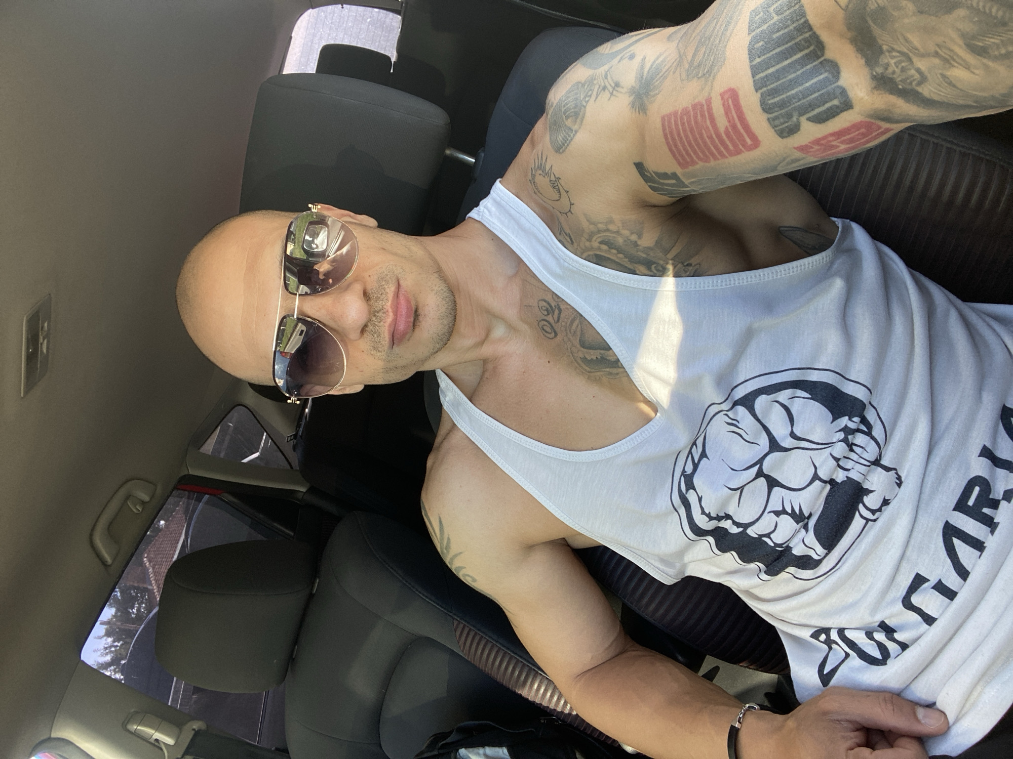 A man with a shaved head and sunglasses is seated in a car. He is wearing a white tank top with a graphic of a clenched fist and the word "Bulgaria" printed on it. The man has numerous tattoos on his arms and chest. The car's interior is visible, including the black leather seats. The location is indicated to be Pleven, Bulgaria, though there is no specific identifying information to support that location visible in the image.