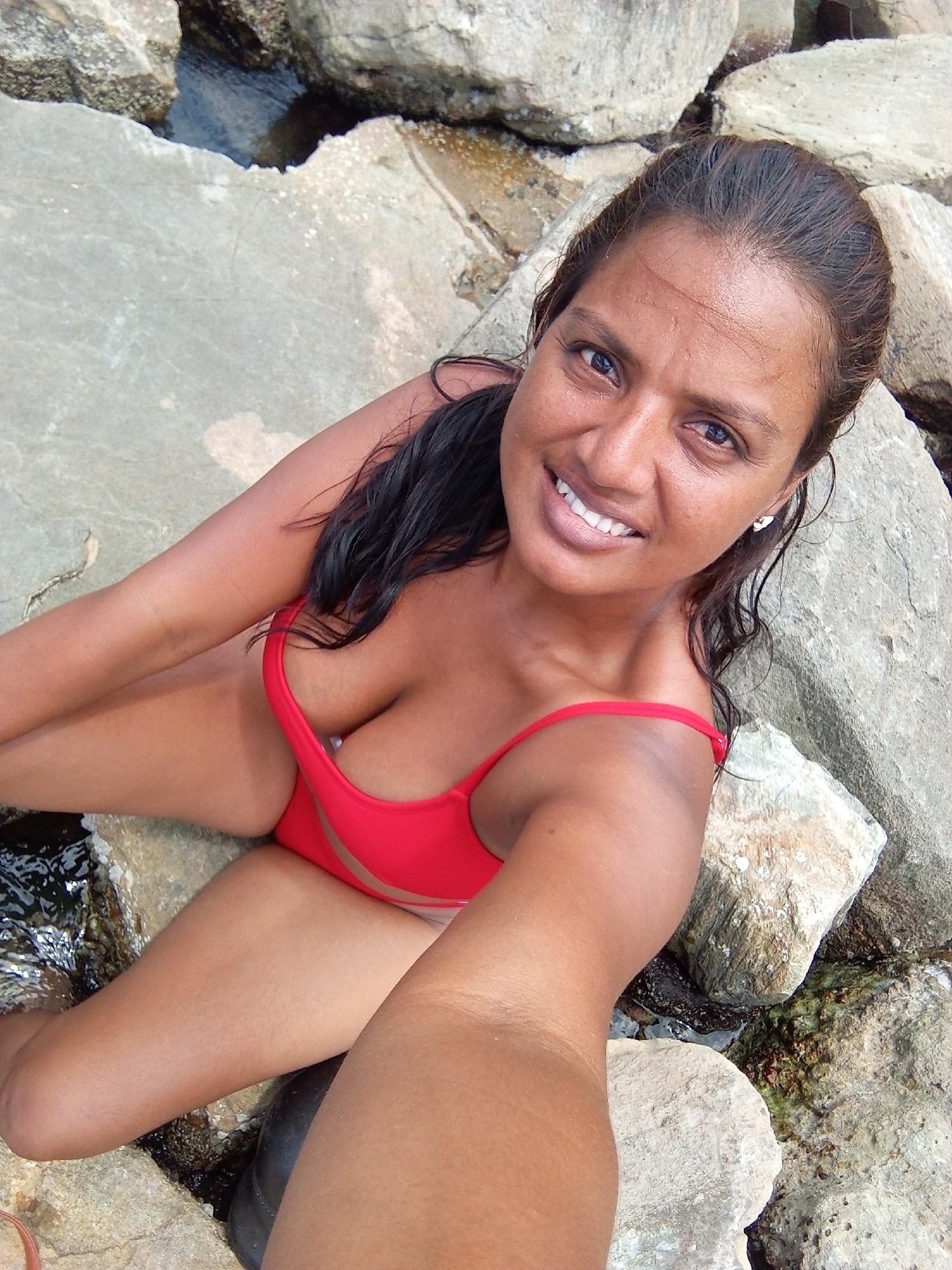 A woman in a red swimsuit is positioned on rocks near a body of water. She is looking at the camera and smiling. Her hair is dark and wet. The rocks are various sizes and appear to be rough. The water is dark and appears shallow in some areas.