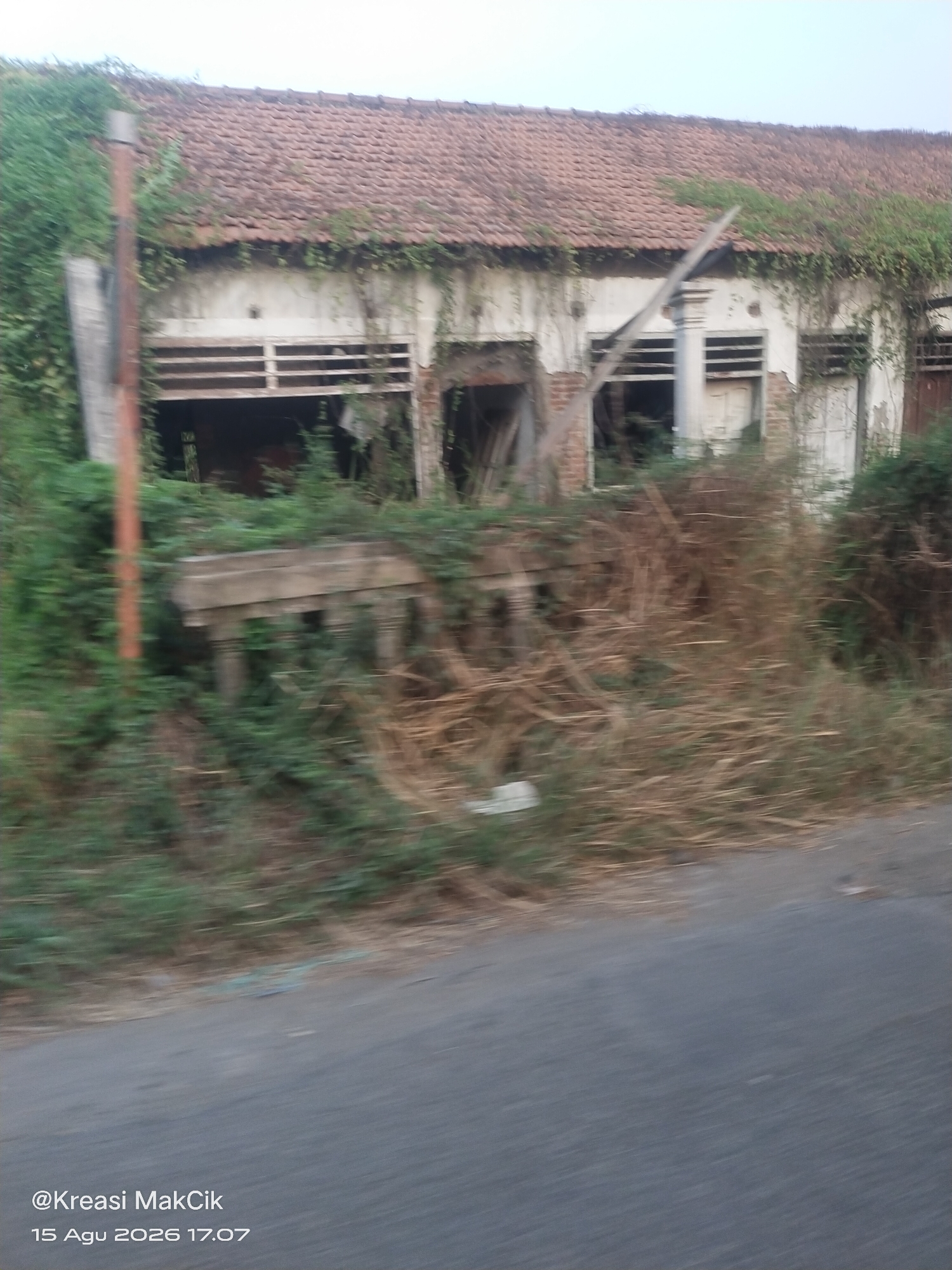The image shows an abandoned building with a red tile roof, covered in vines and greenery. The building appears to be old and in disrepair, with broken windows and crumbling walls. In front of the building, there is a stone structure, possibly a table or a monument, overgrown with vegetation. The scene is framed by a dirt road and a paved road in the foreground. The timestamp in the bottom left corner indicates the image was taken on August 15, 2026, at 5:07 PM, with the watermark "@Kreasi MakCik". The location is identified as Kletek, Indonesia. The image is blurred, suggesting it was taken from a moving vehicle.