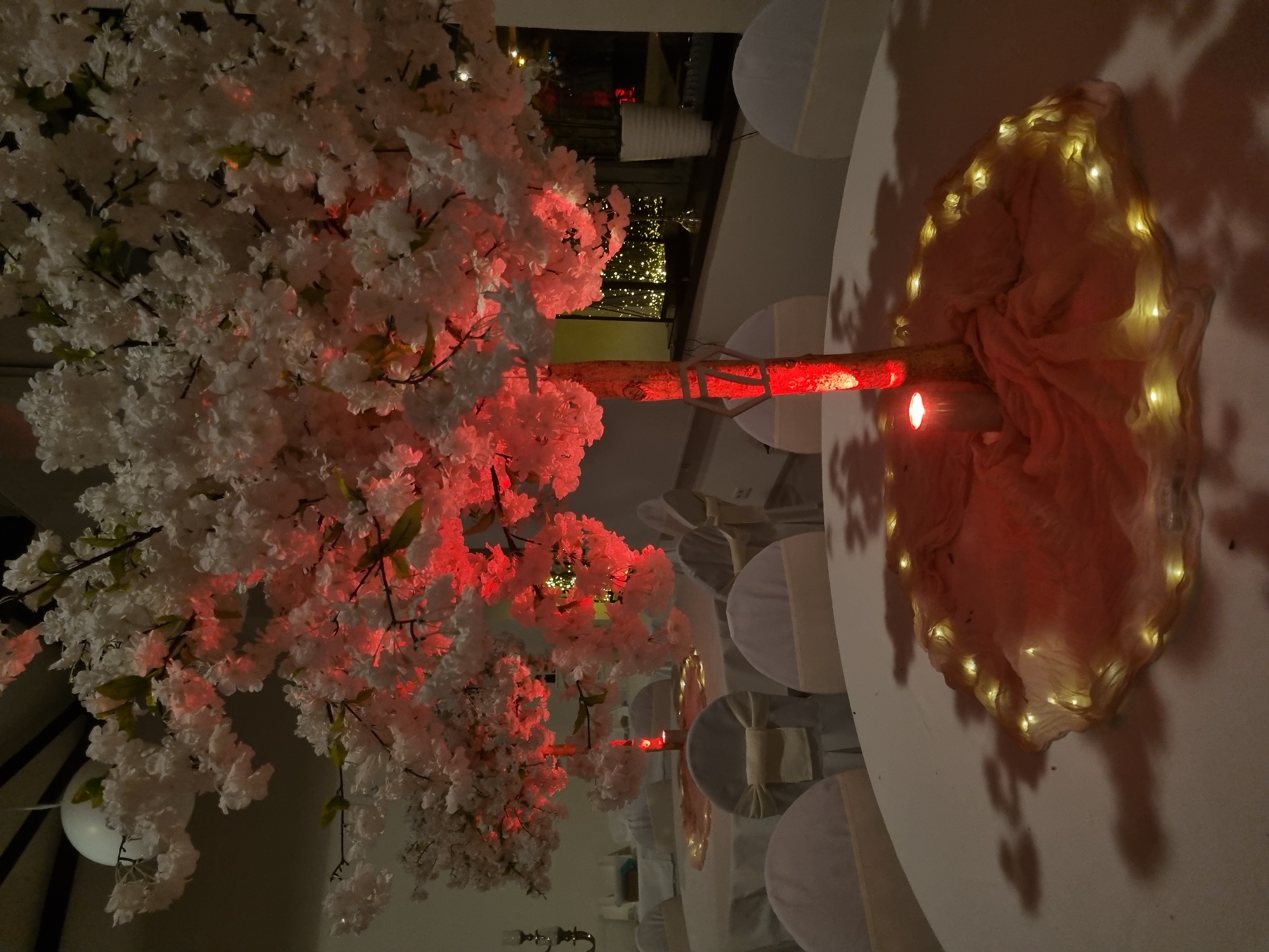 The image shows a wedding reception in Saarbrücken, Germany. A white artificial tree with red lighting serves as a centerpiece on a round table. The base of the tree is wrapped in a light-colored, draped fabric illuminated with small fairy lights. A lit candle sits at the center of the base. Surrounding the table are white chairs with matching white chair covers and bows. The background shows a blurred view of the event space with lights and other tables.