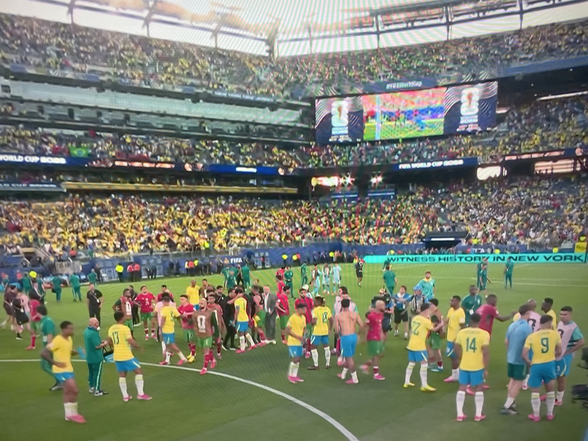 A wide shot captures a football (soccer) stadium filled with spectators. The field is occupied by numerous football players in distinct team uniforms. Some players are gathered in groups, possibly interacting with each other or coaches. The stands are packed with a predominantly yellow-clad crowd. Large screens are visible above the stands, displaying images of the game or players. A banner across the lower part of the stadium reads "WITNESS HISTORY IN NEW YORK". The overall atmosphere appears to be that of a professional sporting event. The location is not the city of Tbilisi, Georgia, but likely a stadium in New York as indicated by the banner.